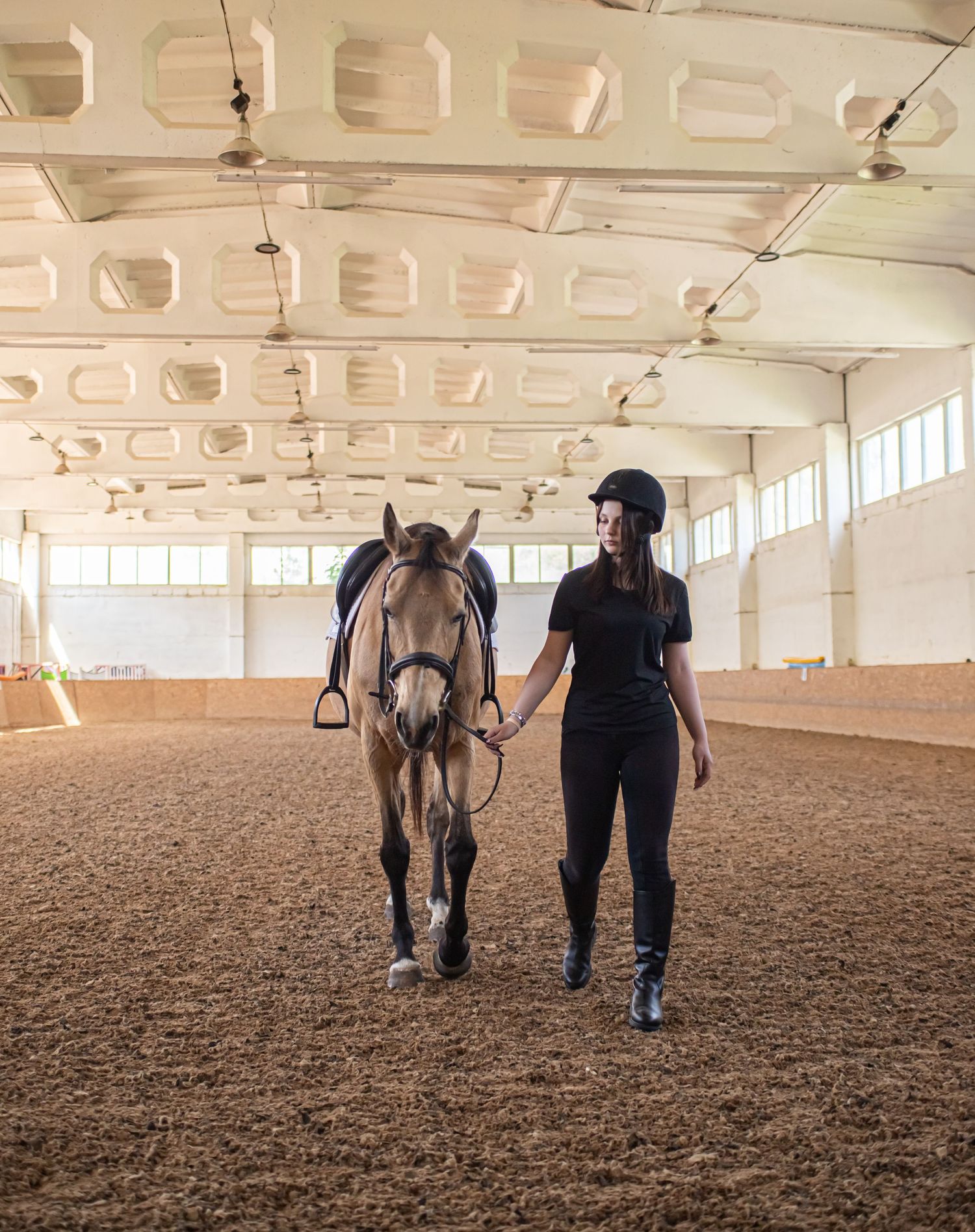 Female equestrian in black riding attire leading a buckskin horse in an indoor arena.