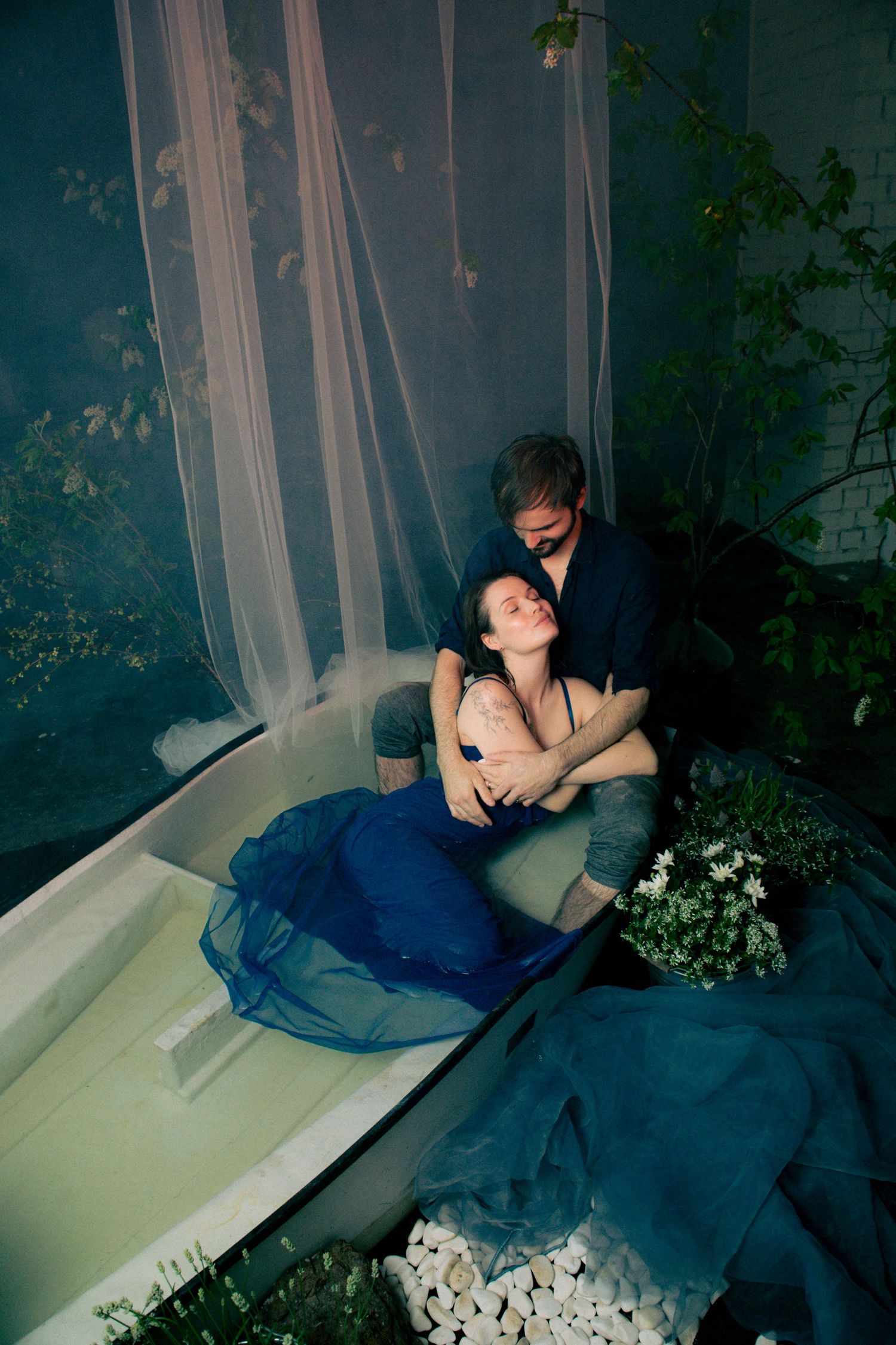 Romantic couple embracing in a bathtub surrounded by lush greenery, sheer curtains, and white flowers in moody blue lighting.