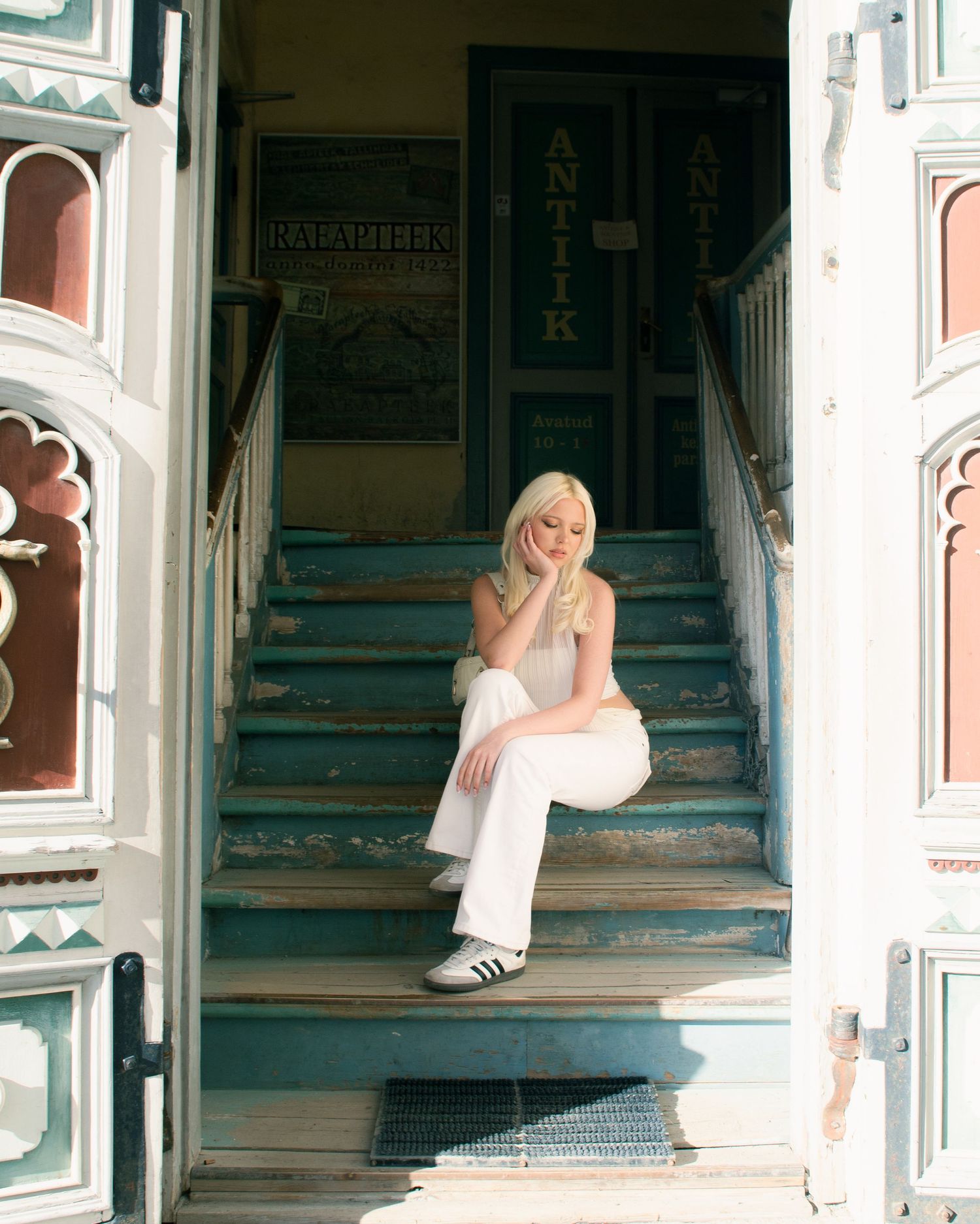 Blonde woman in white outfit sitting on teal vintage steps between ornate doors of an antique shop.
