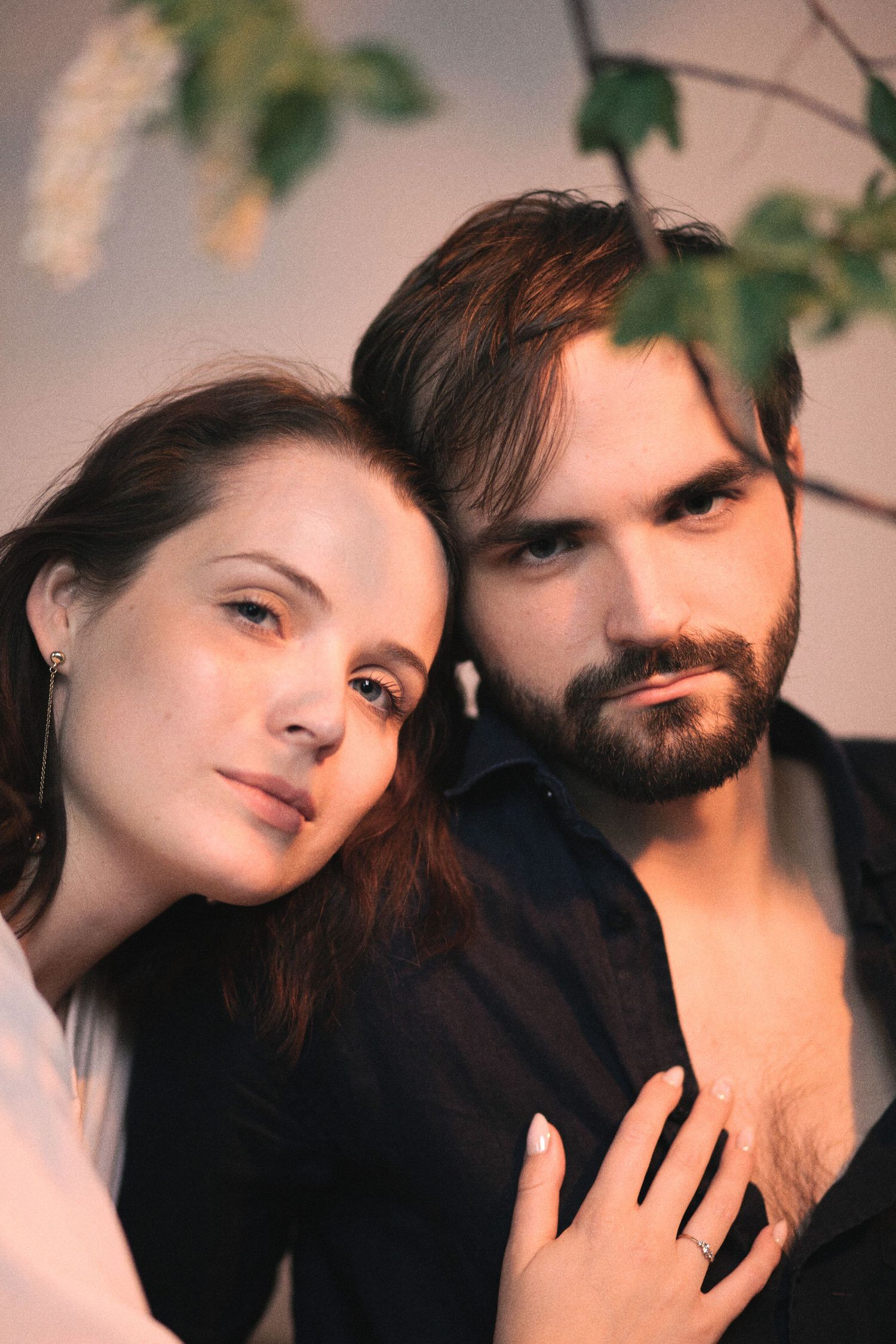 A young couple poses intimately, woman leaning on bearded mans shoulder, surrounded by green leaves indoors.