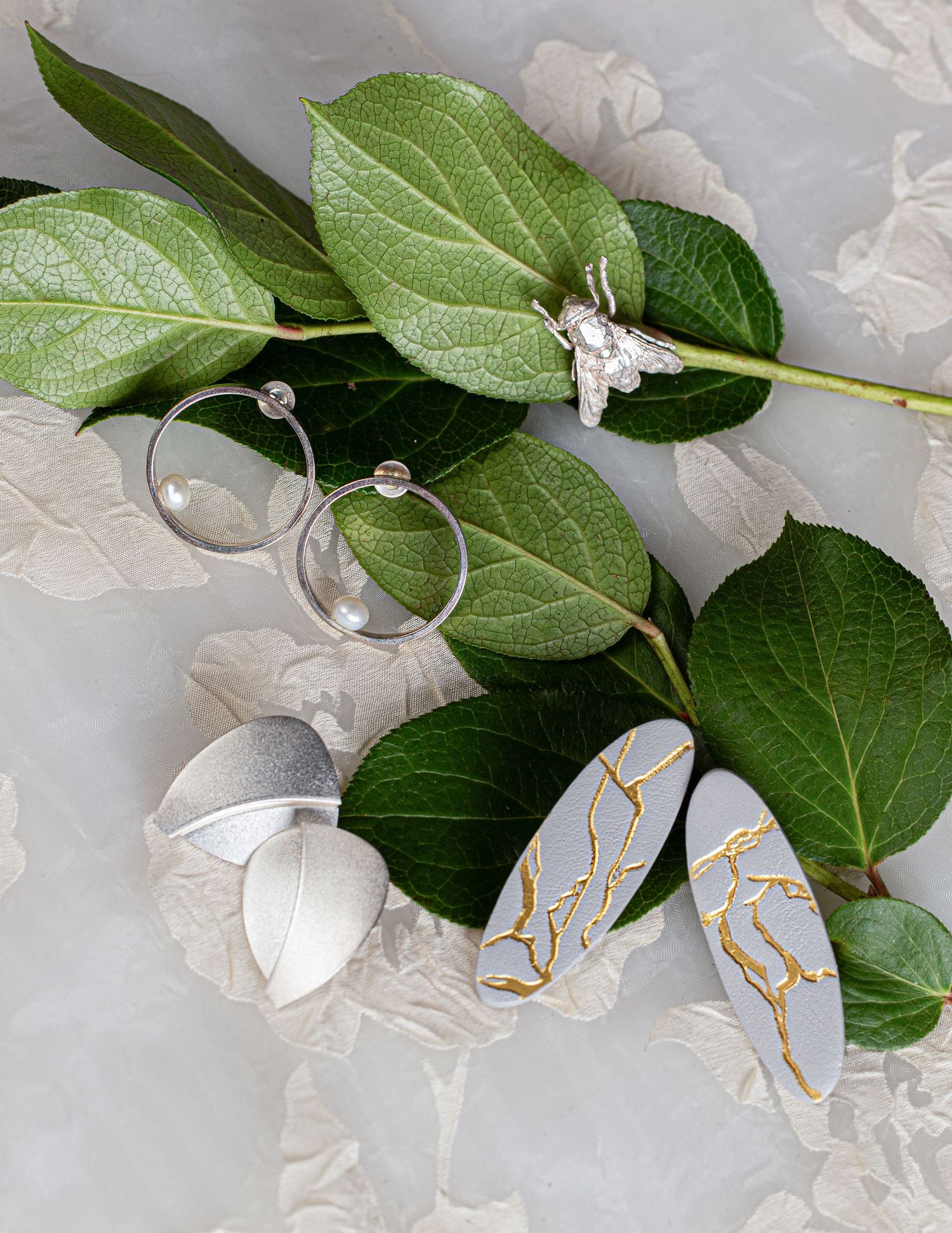 Silver hoop earrings, crystal ring, and gray kintsugi-style earrings with gold accents displayed on green leaves.