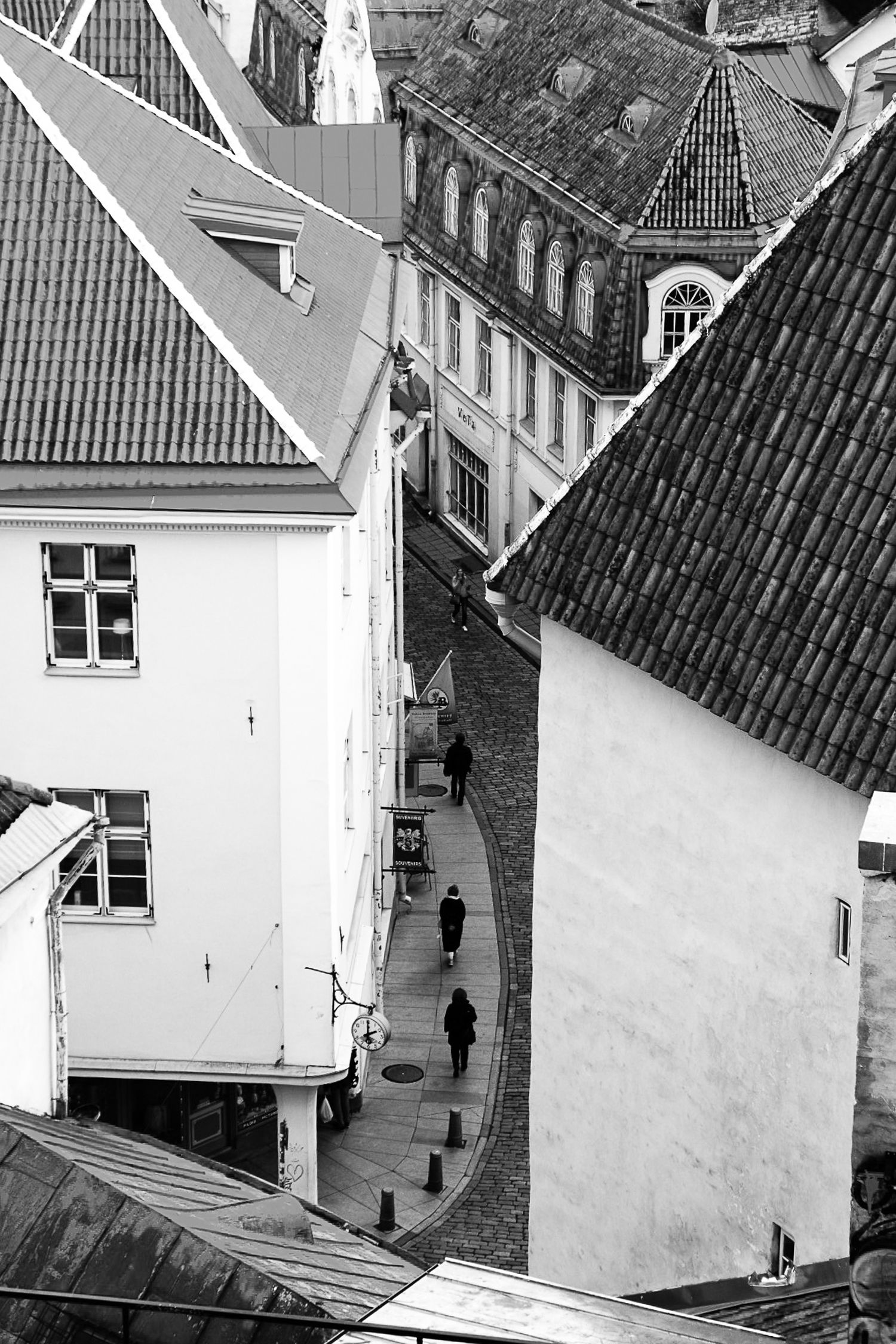 Aerial black and white view of narrow cobblestone street between historic European buildings with tiled roofs.