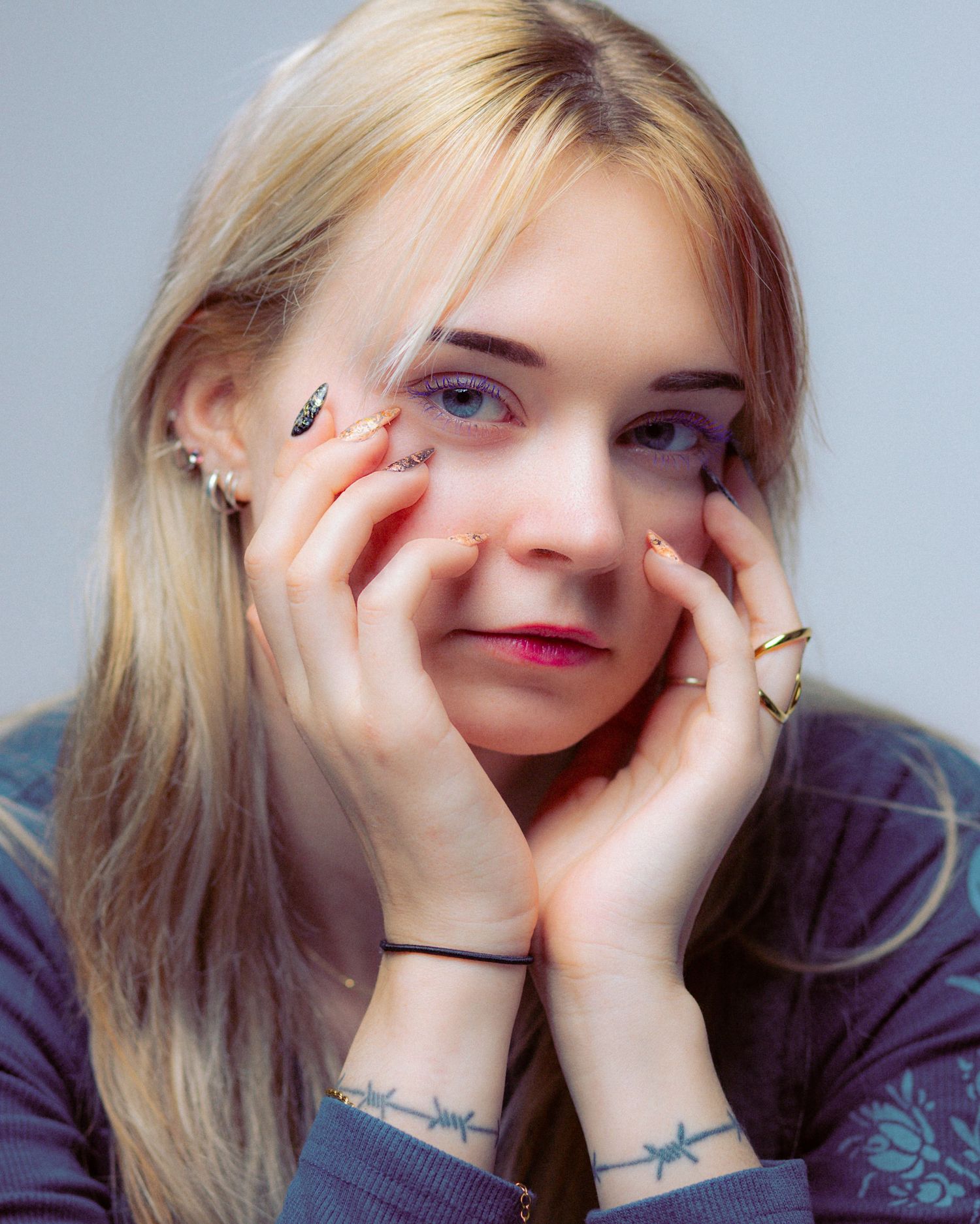 Young blonde woman resting face in hands, wearing rings and barbed wire wrist tattoos, with colorful nail art.