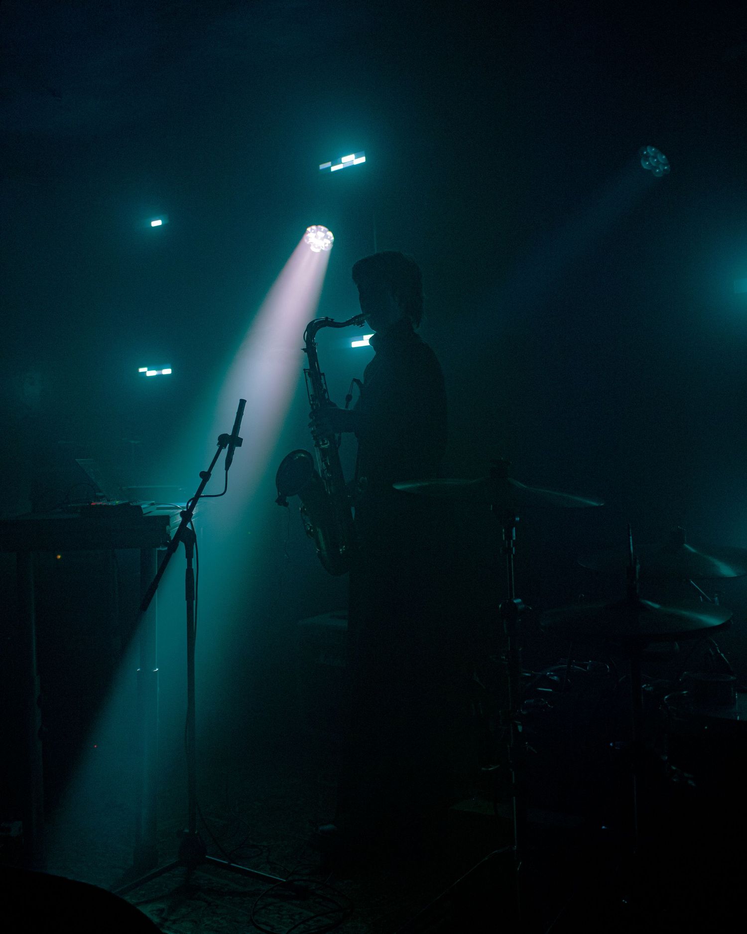 Silhouetted saxophonist performing on stage under a dramatic spotlight with teal-toned atmospheric haze at a live concert.