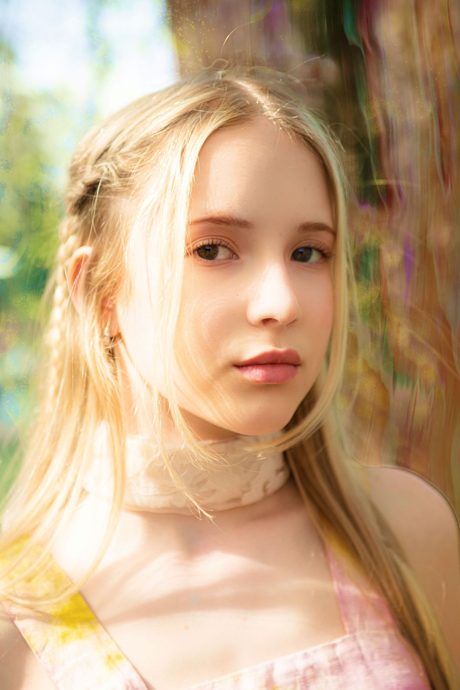 Young blonde girl with long hair posing outdoors in soft natural sunlight with colorful bokeh background.