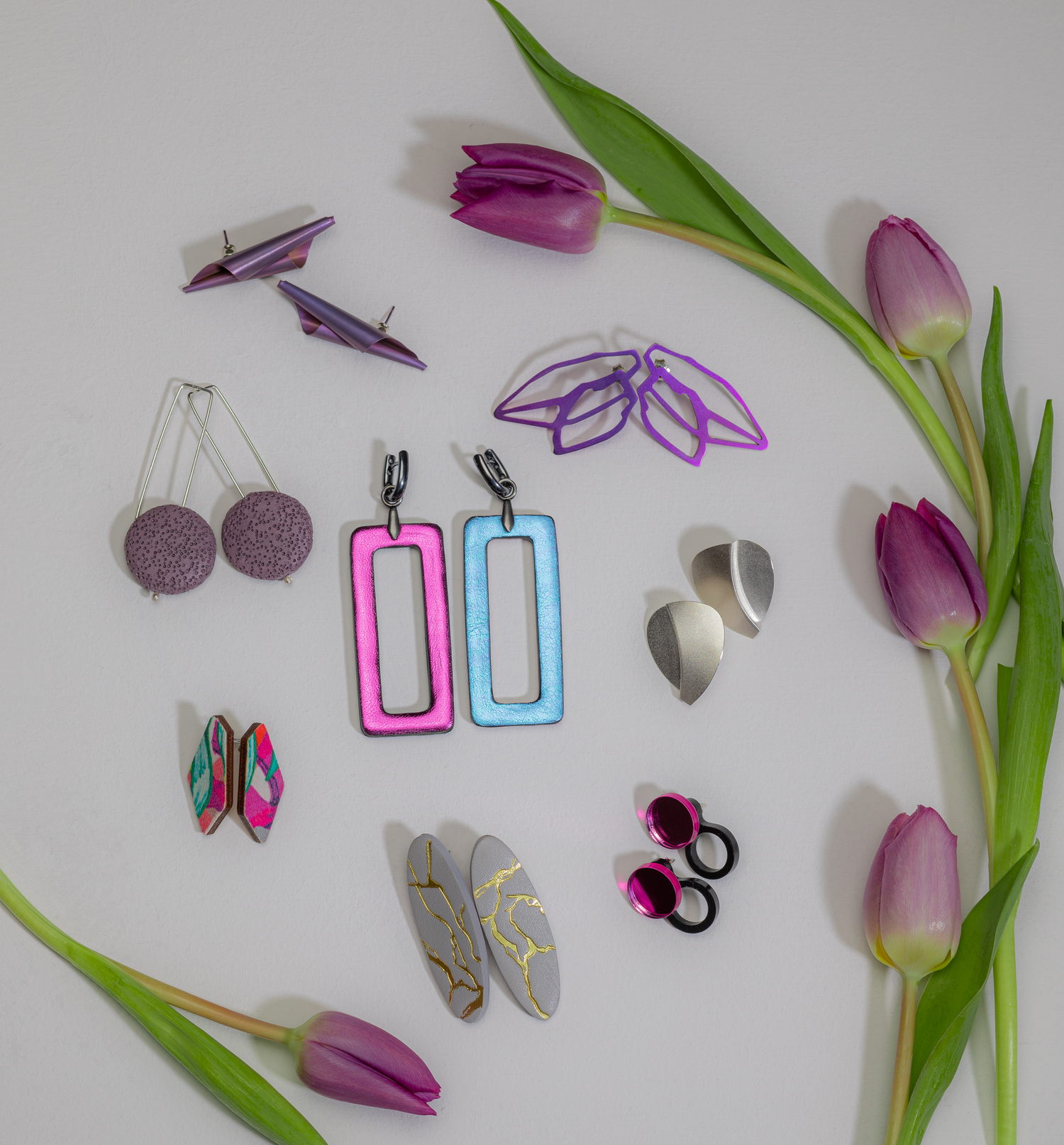 Colorful collection of handmade earrings in various shapes displayed with purple tulips on white background.