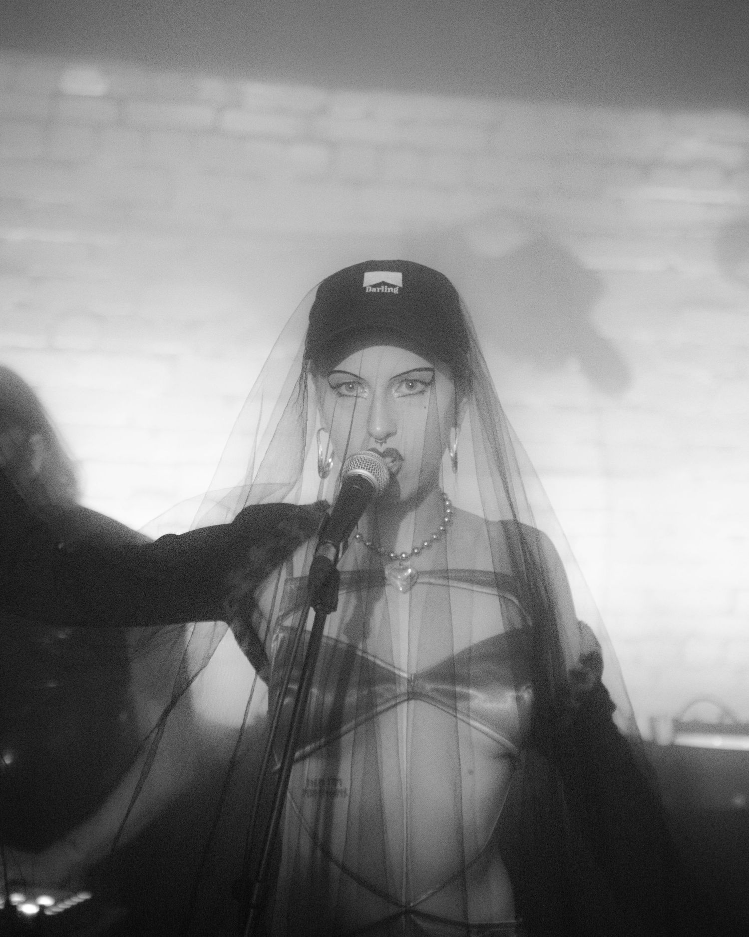 Black and white close-up of a female singer wearing a veil and cap performing at a microphone.
