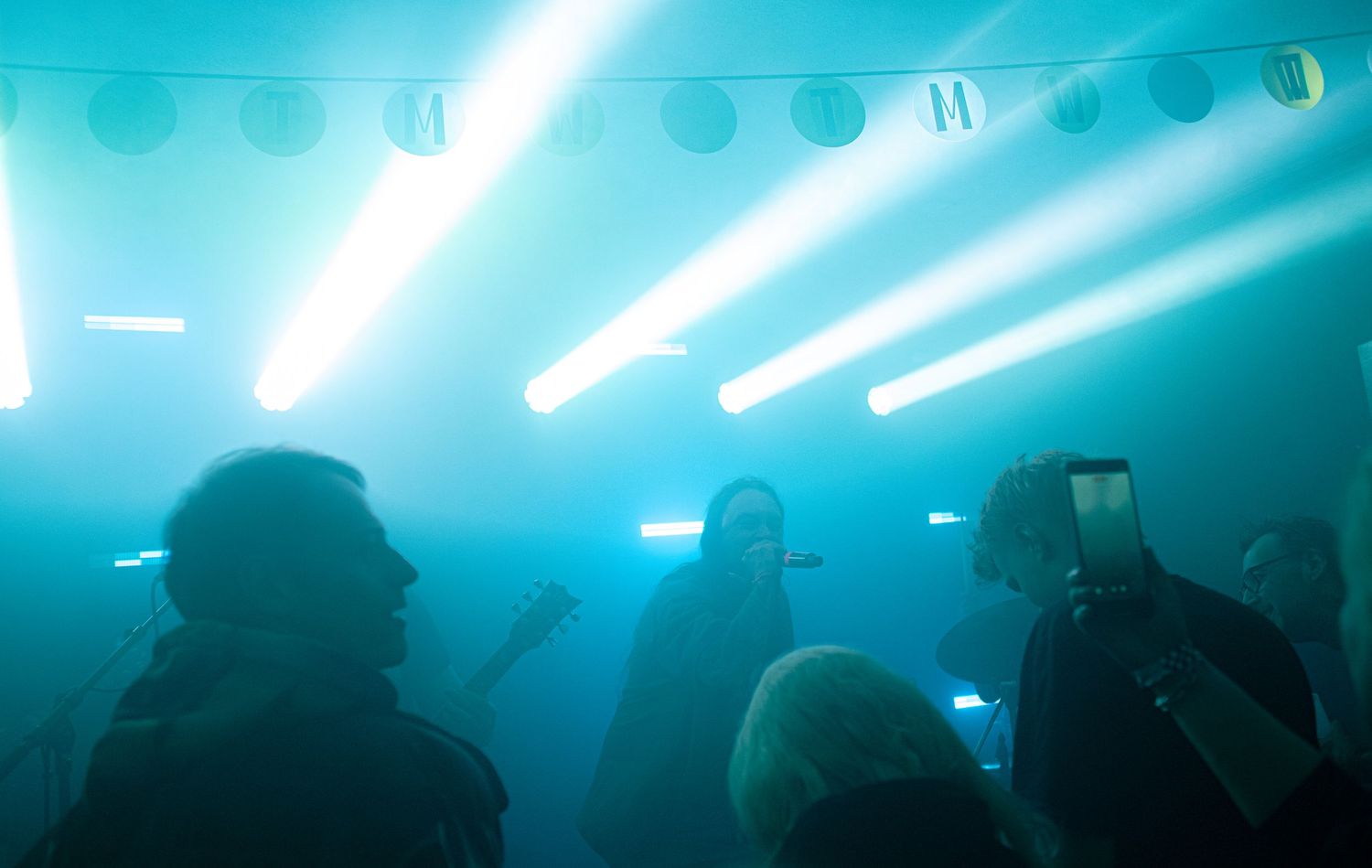 Live concert scene with dramatic cyan stage lighting beams cutting through smoke above silhouetted crowd members.