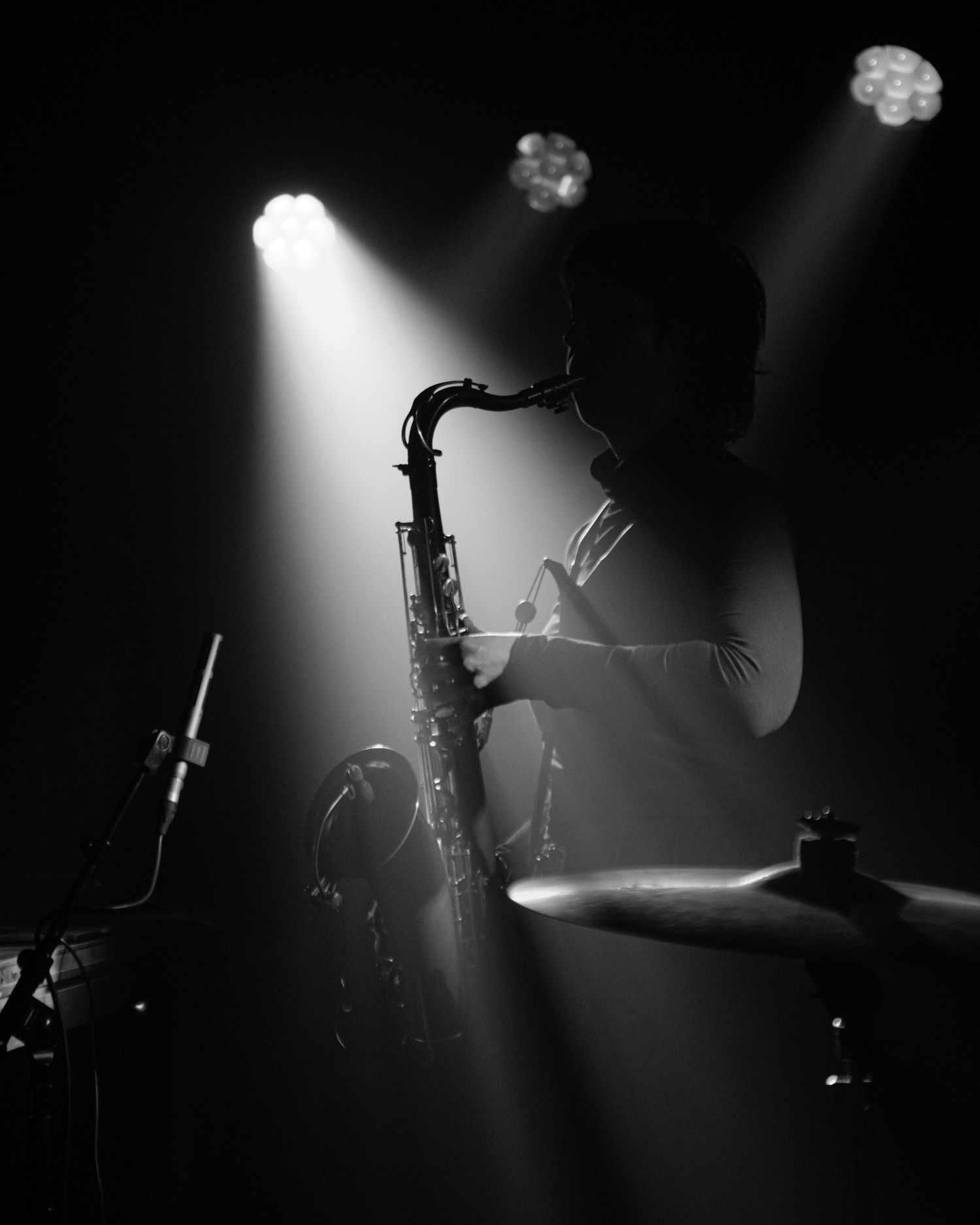 Black and white photo of a saxophonist performing on stage under dramatic spotlights with drum cymbals visible in foreground.