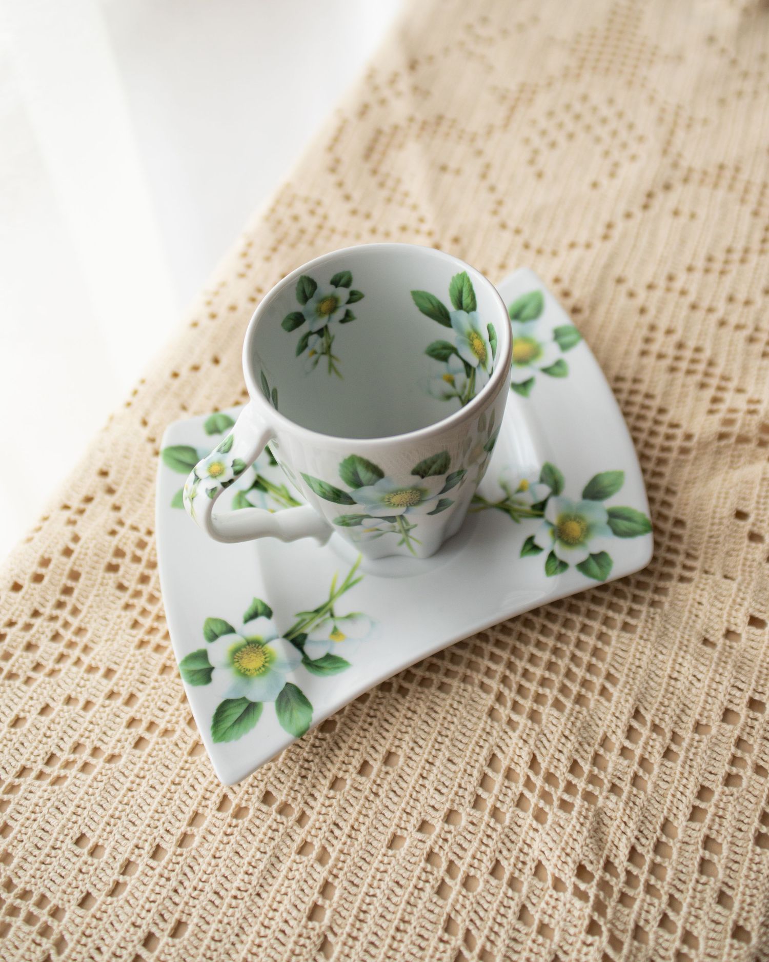 White porcelain tea cup and square saucer with green floral pattern on lace tablecloth.