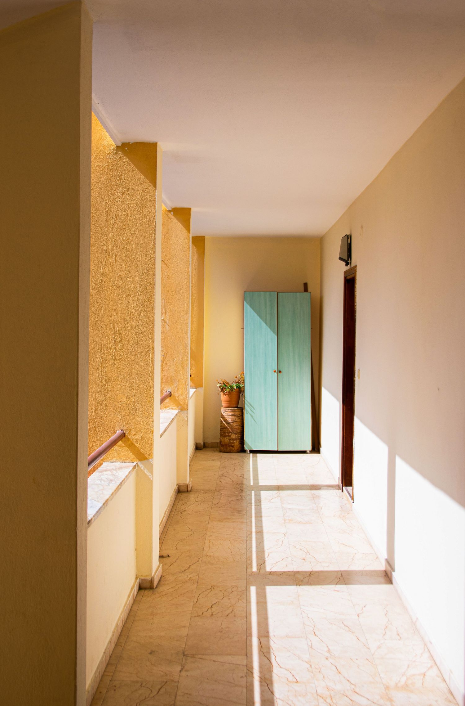 Sunlit corridor with yellow walls, marble floor, and a turquoise cabinet at the end.