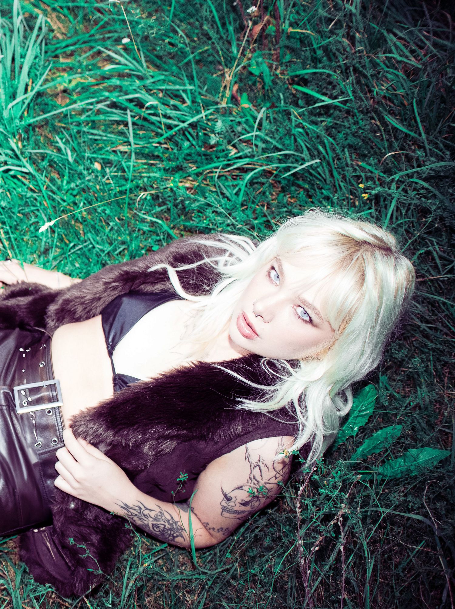 Tattooed platinum blonde woman in a dark fur vest lying in vibrant green grass, smiling softly at the camera.