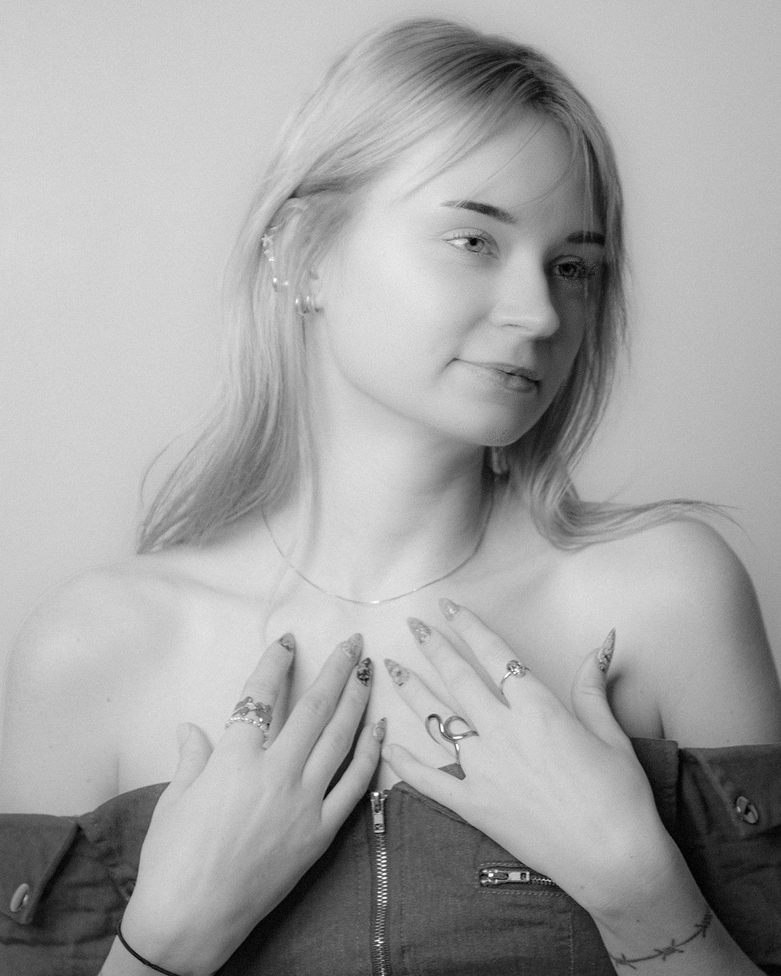 Black and white portrait of a young blonde woman wearing rings, necklace, and off-shoulder top, posing gracefully.