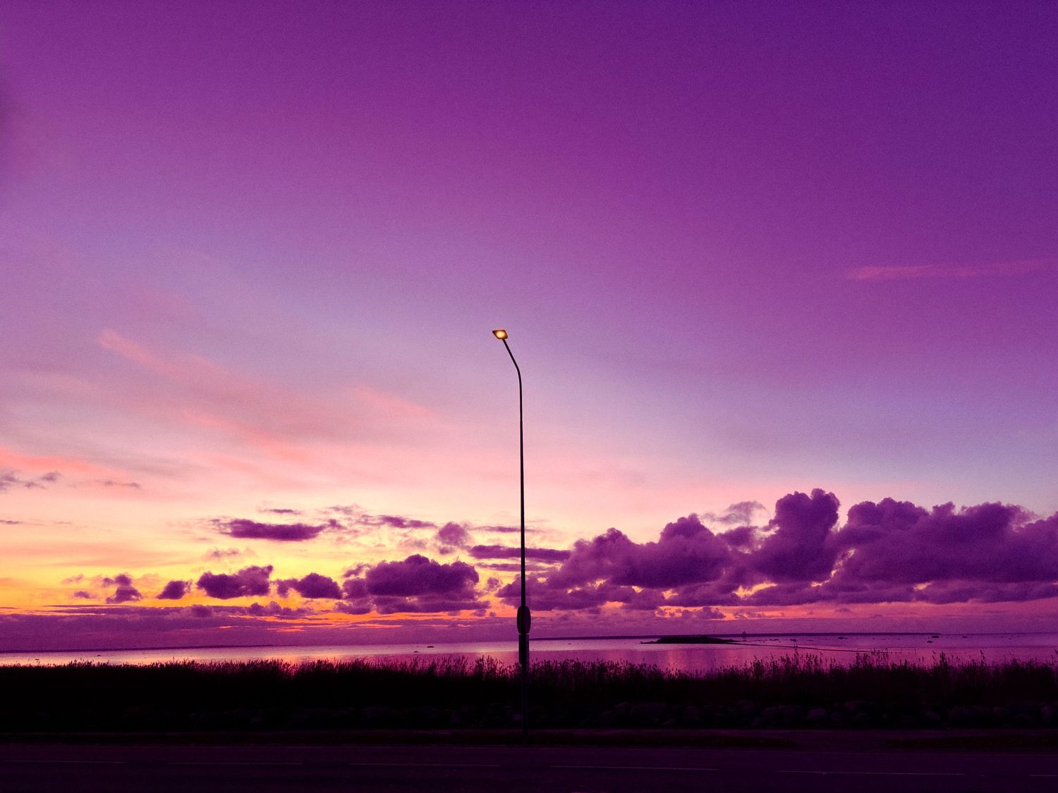 Vibrant purple sunset sky with dramatic clouds over water and a lone street lamp silhouette.