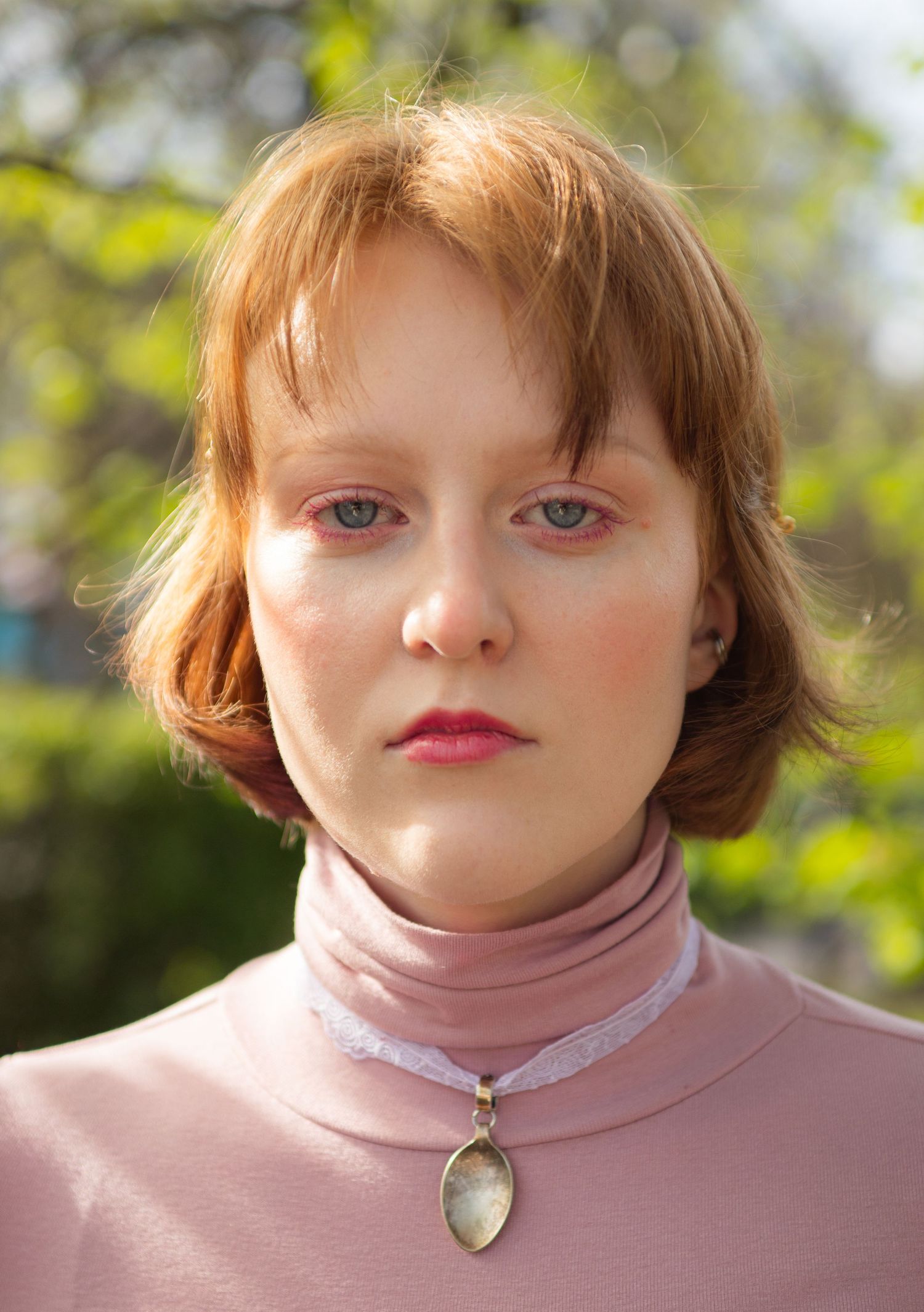 Young woman with red hair and blue eyes wearing a pink turtleneck and pendant necklace outdoors.