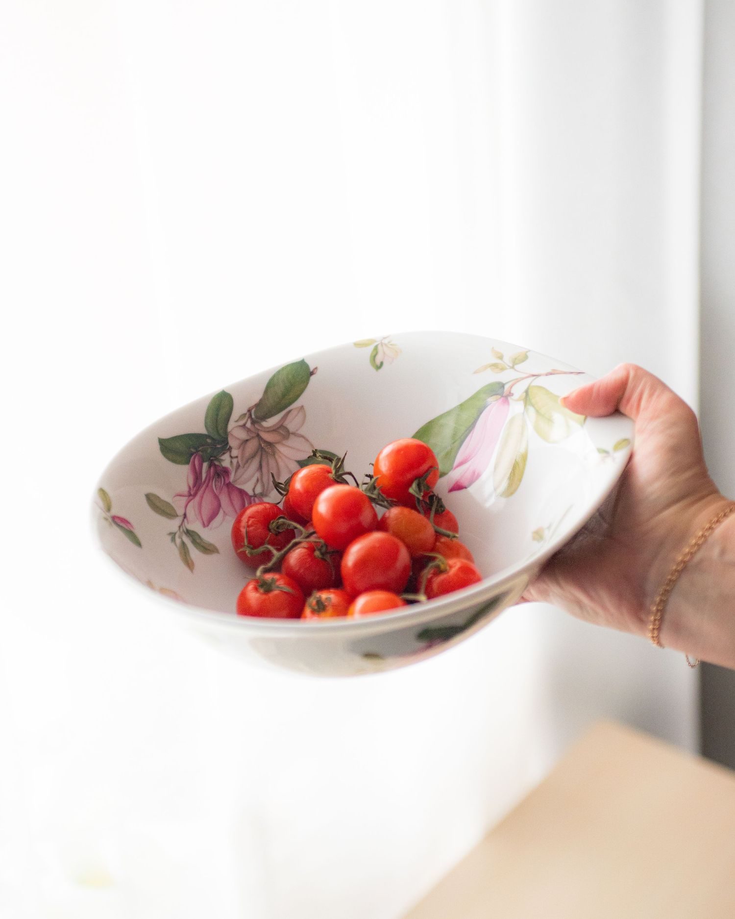 Hand holding a white floral ceramic bowl filled with fresh red cherry tomatoes on the vine.