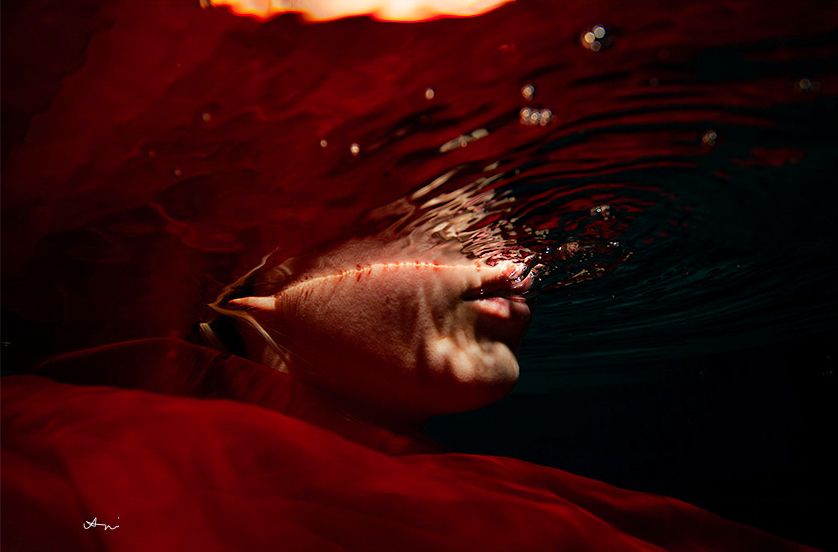 Person submerged underwater surrounded by flowing red fabric, face partially above water in dramatic artistic photography.