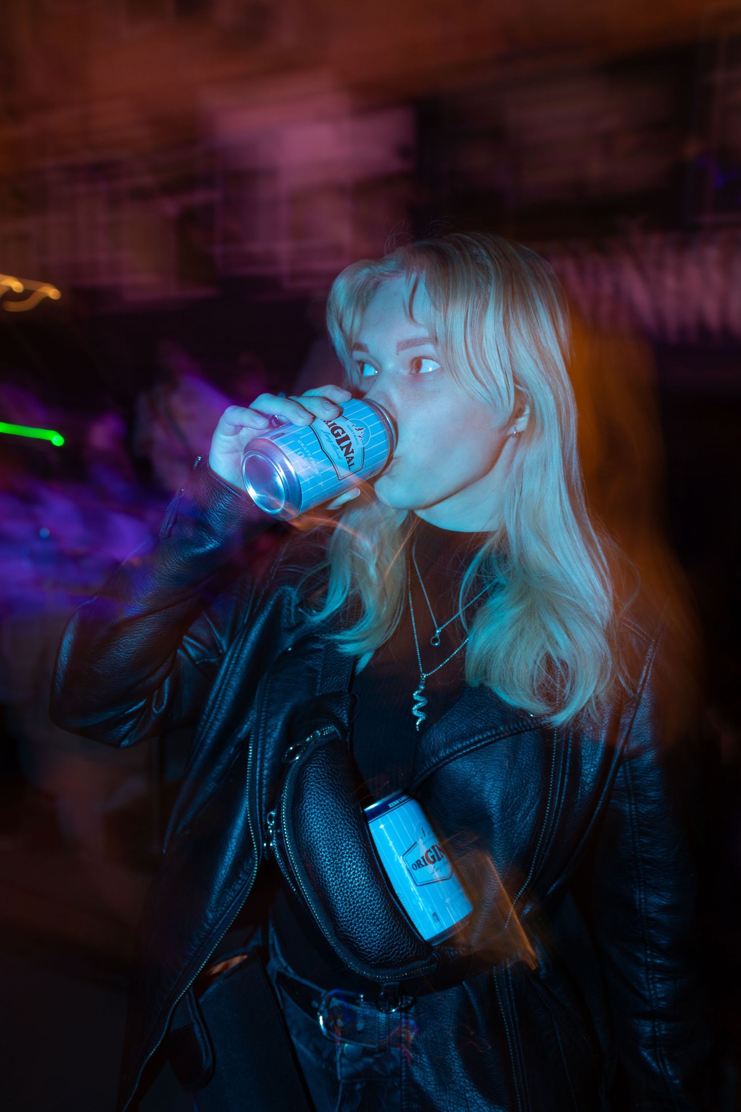 Blonde woman in leather jacket drinking from a can under colorful neon lights at night event.