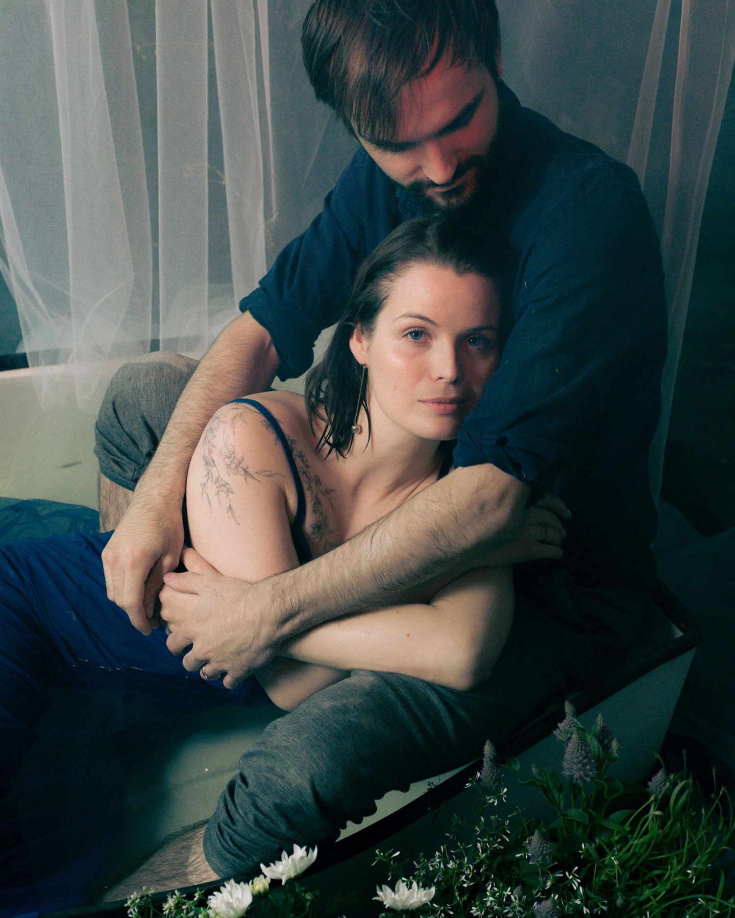 A man and woman in an intimate embrace, sitting together with floral tattoos visible on her arm.