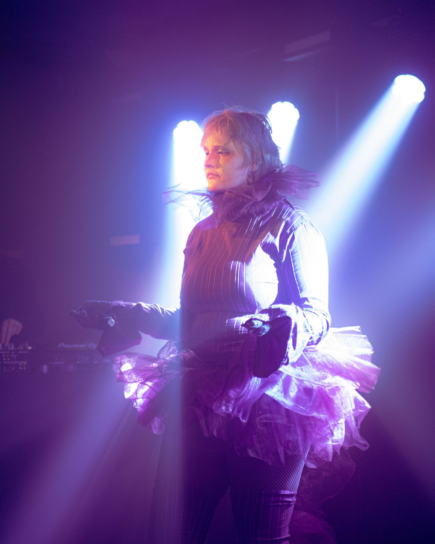 Performer in purple tutu and dark jacket under dramatic stage lighting with colorful beams at a live concert.