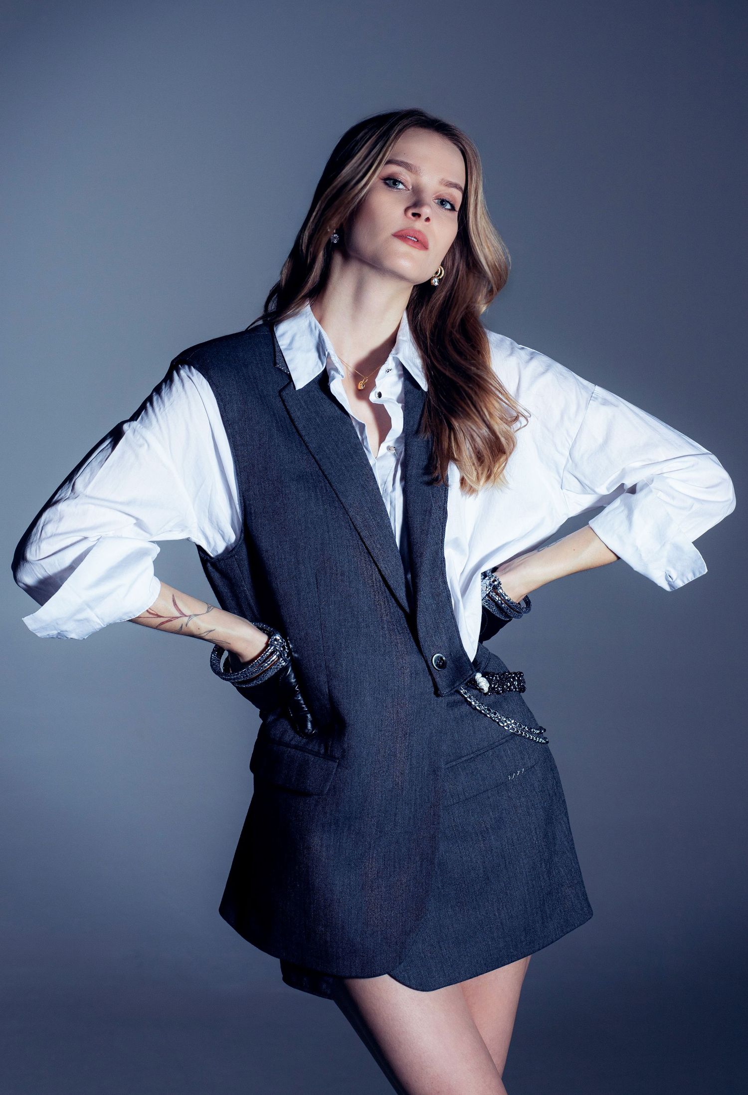 Woman posing confidently in a white shirt and dark gray sleeveless blazer dress with chain details.