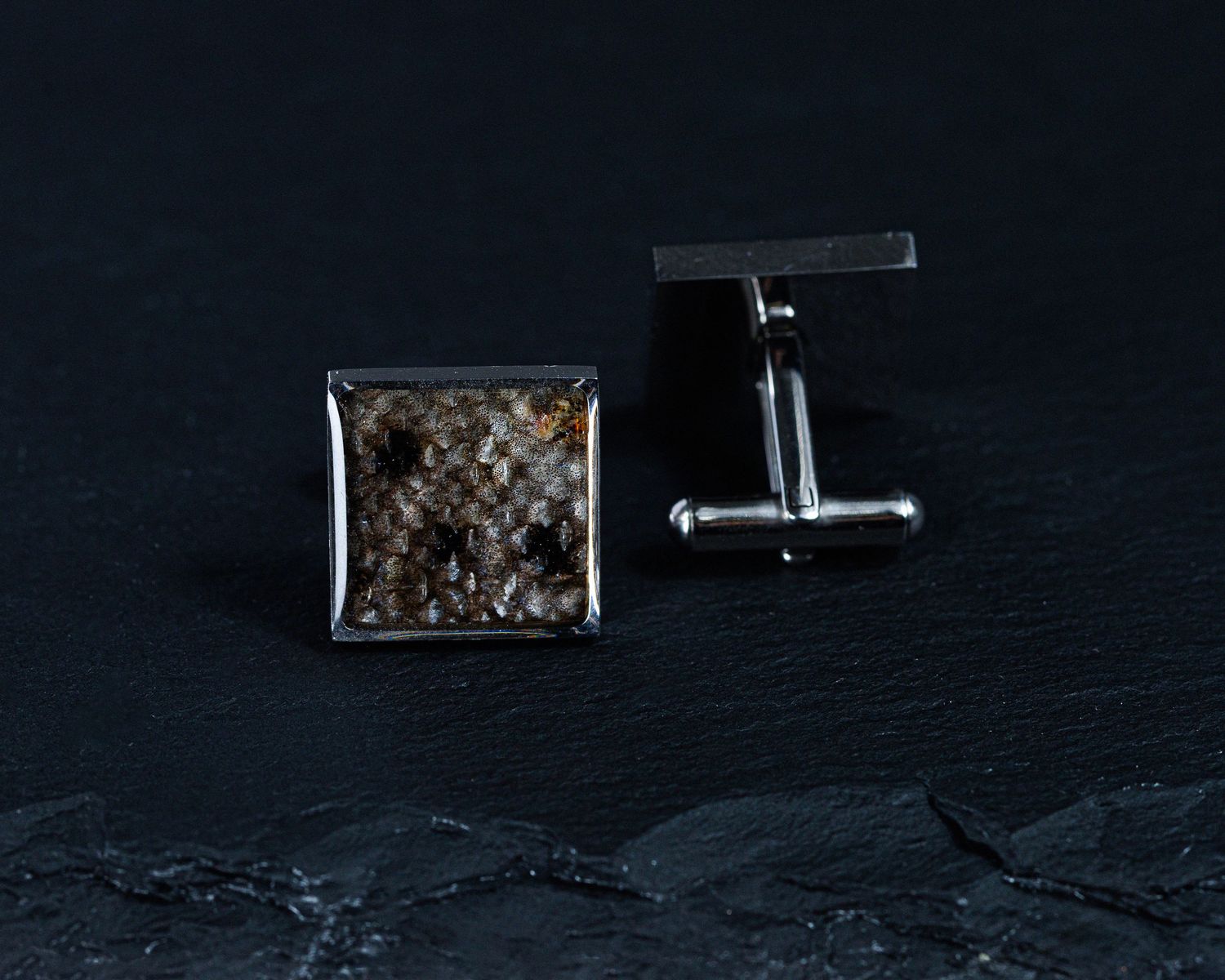 Square silver cufflinks with dark brown textured inlay on a black stone surface.