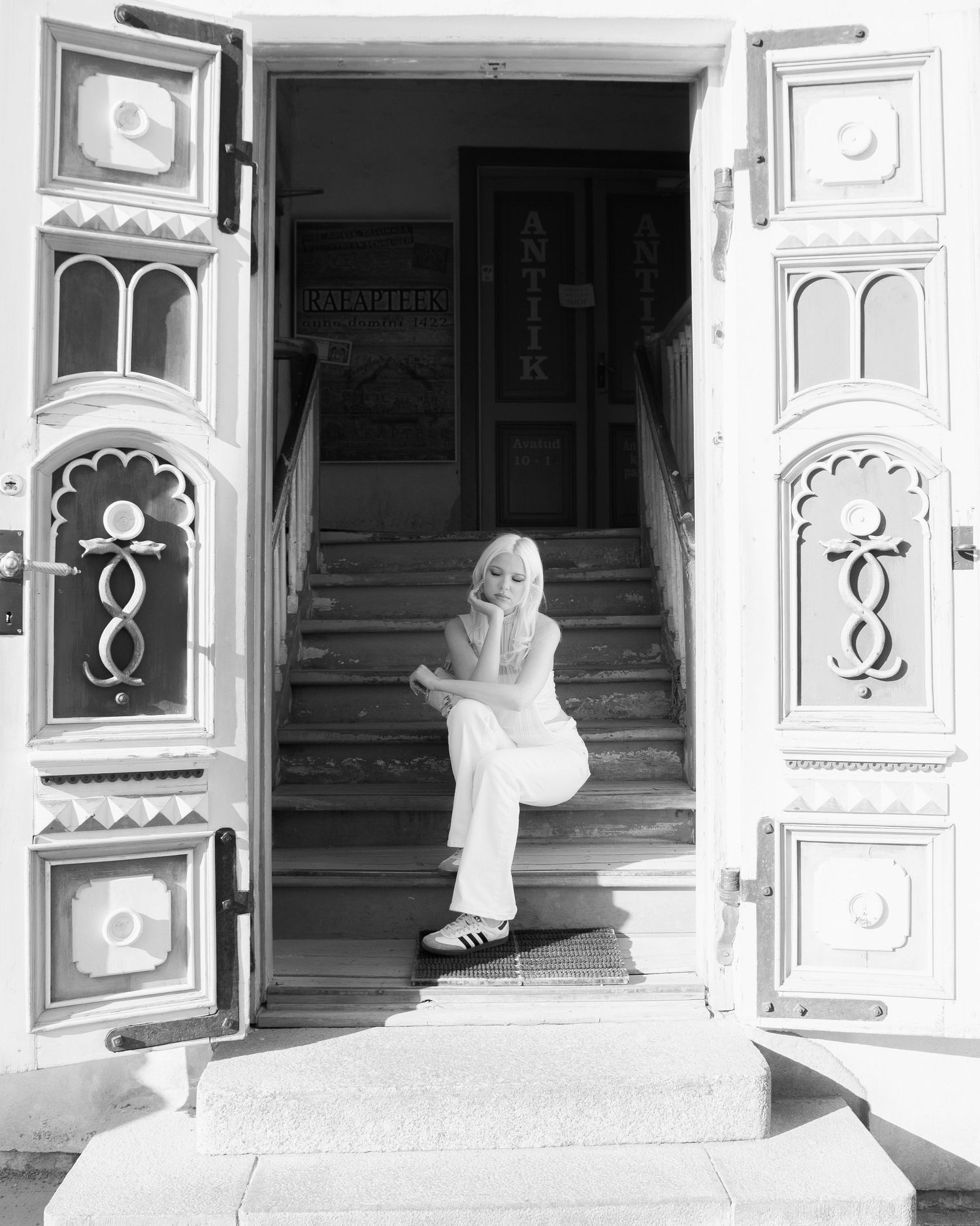 Black and white photo of a blonde woman in white outfit sitting on steps between ornate decorated doors.