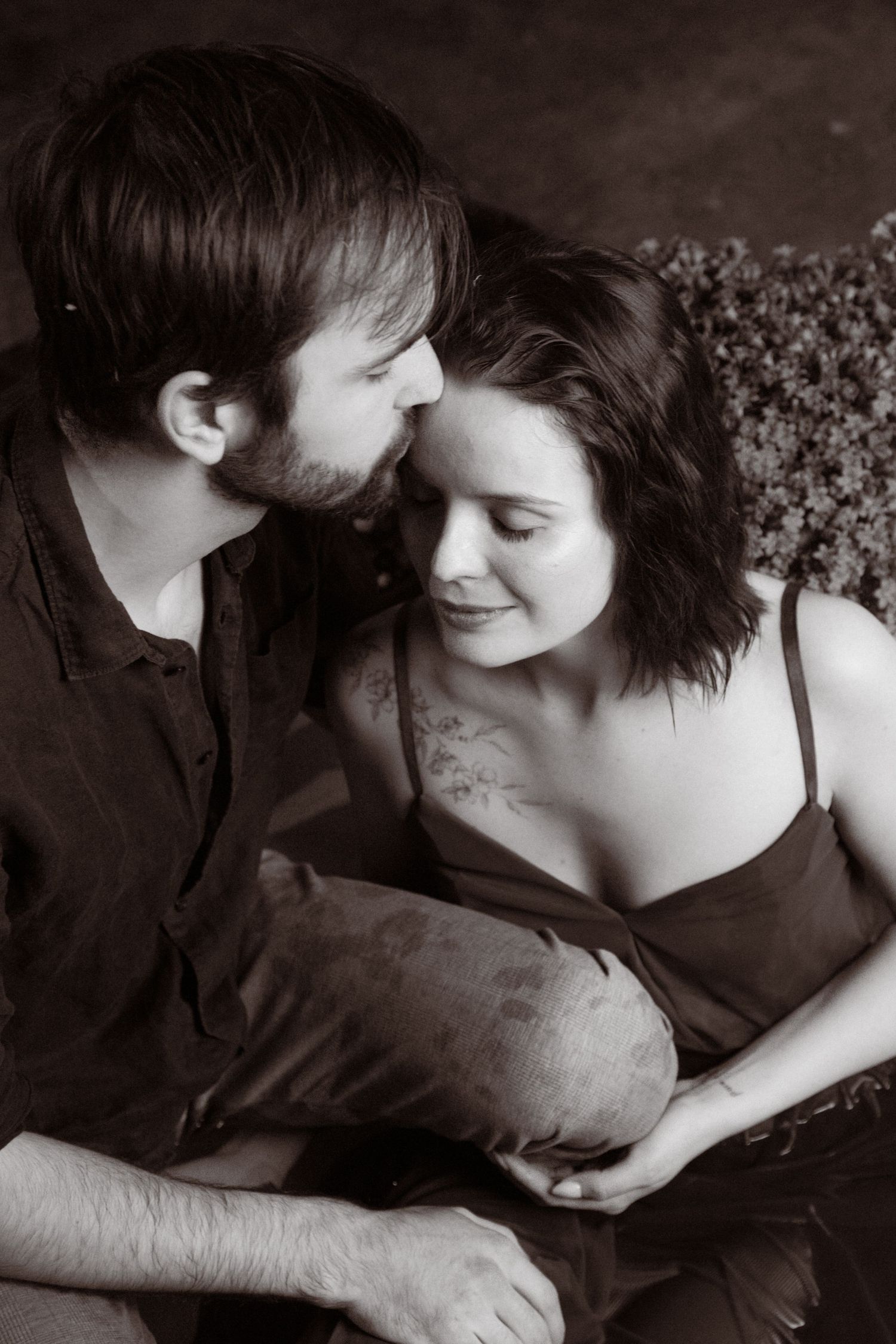Black and white photo of a romantic couple sitting closely together, man leaning in to kiss woman's cheek tenderly.