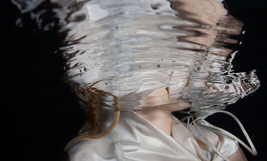 Person submerged underwater wearing a white dress, with rippling water distorting their face and hair.