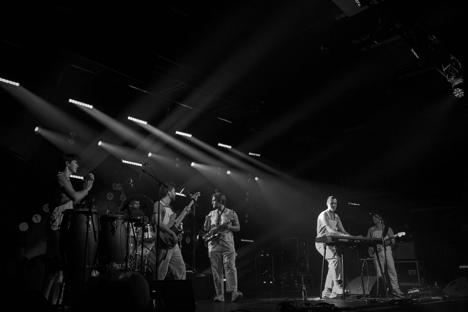 Black and white concert photo featuring a band performing on stage with dramatic light beams overhead.