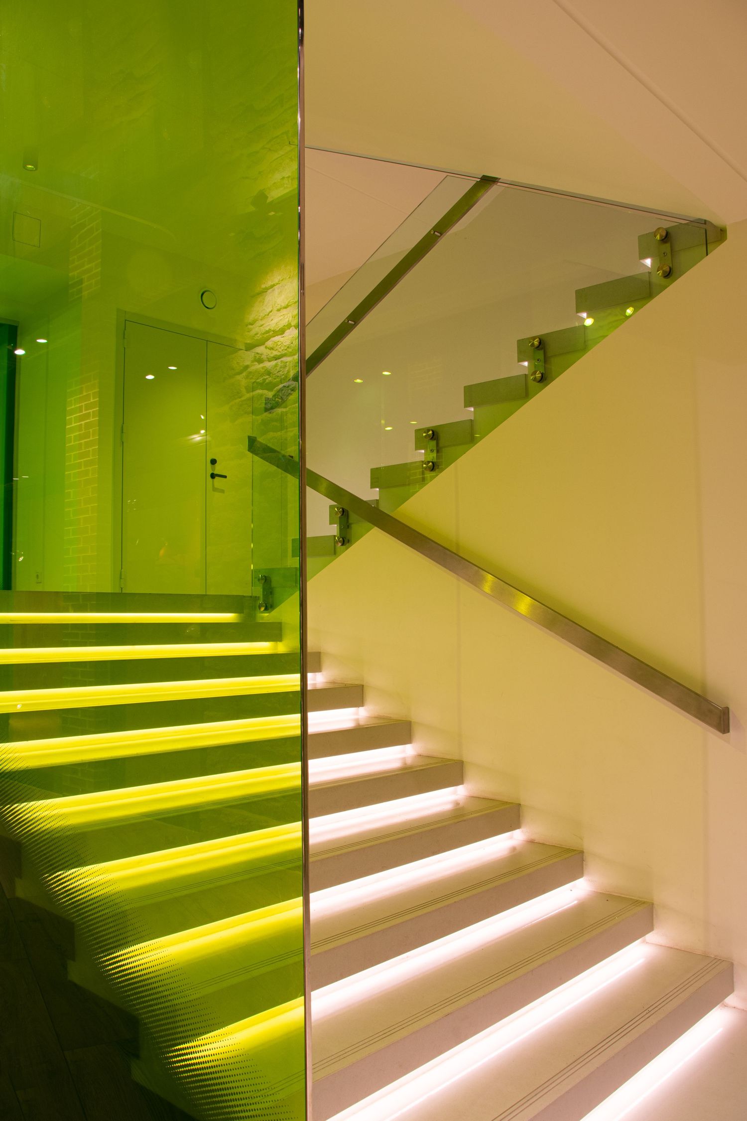 Modern staircase with green LED lighting on steps, glass railing, and reflective green wall panel beside it.
