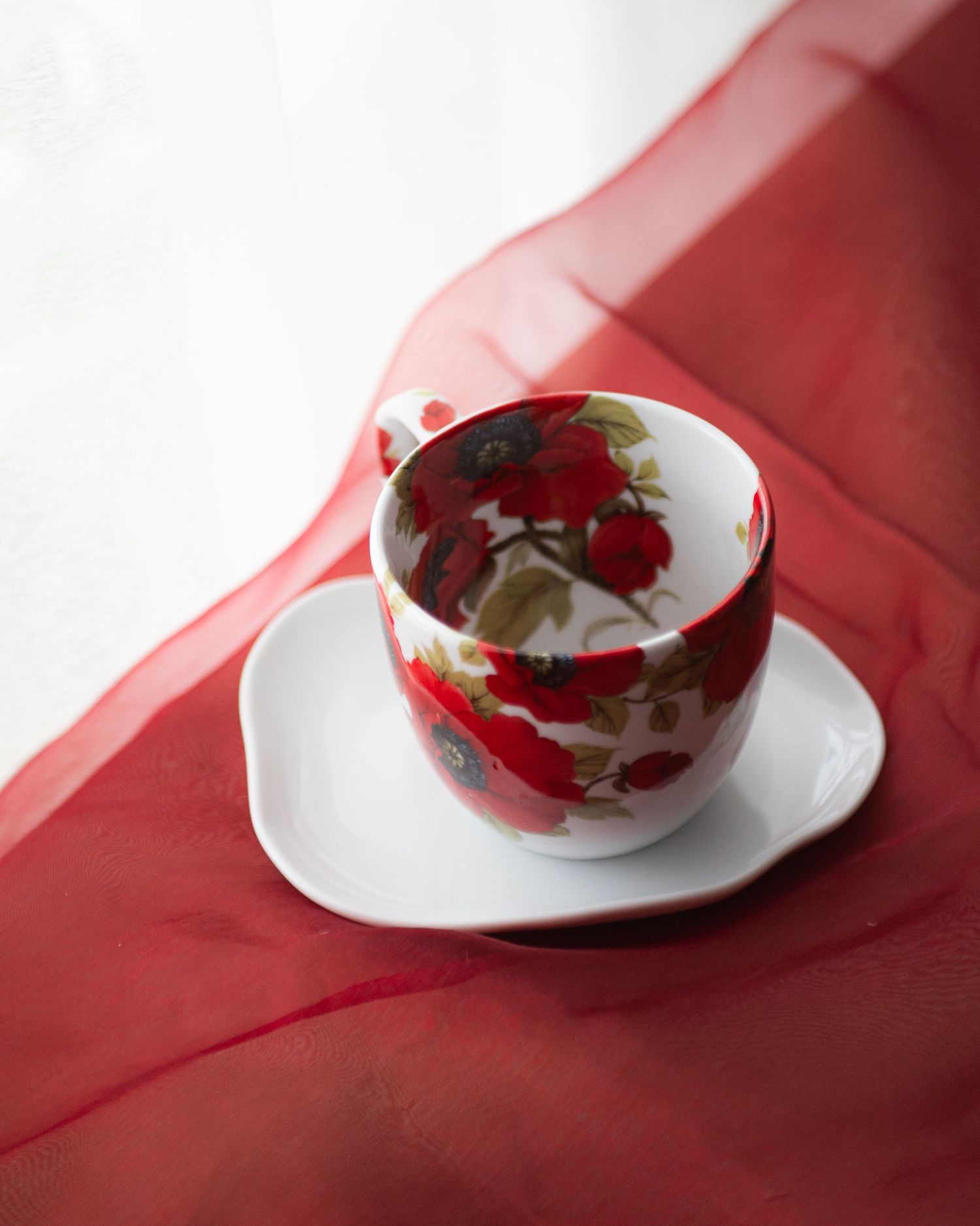 White porcelain teacup with red floral pattern on saucer, draped with sheer red fabric near window.