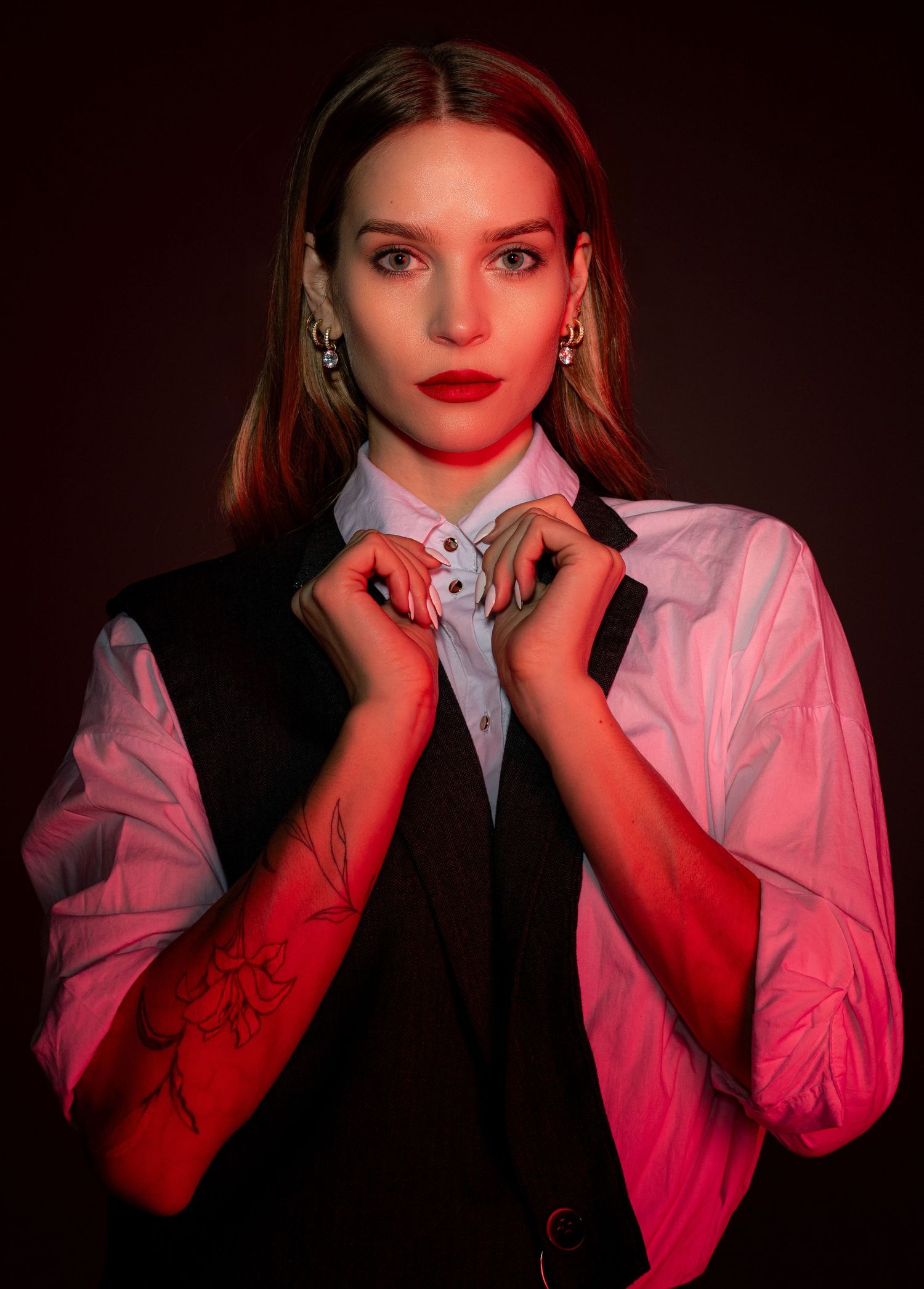 Close-up of woman in black vest and white shirt with rose tattoo under warm red studio lighting.