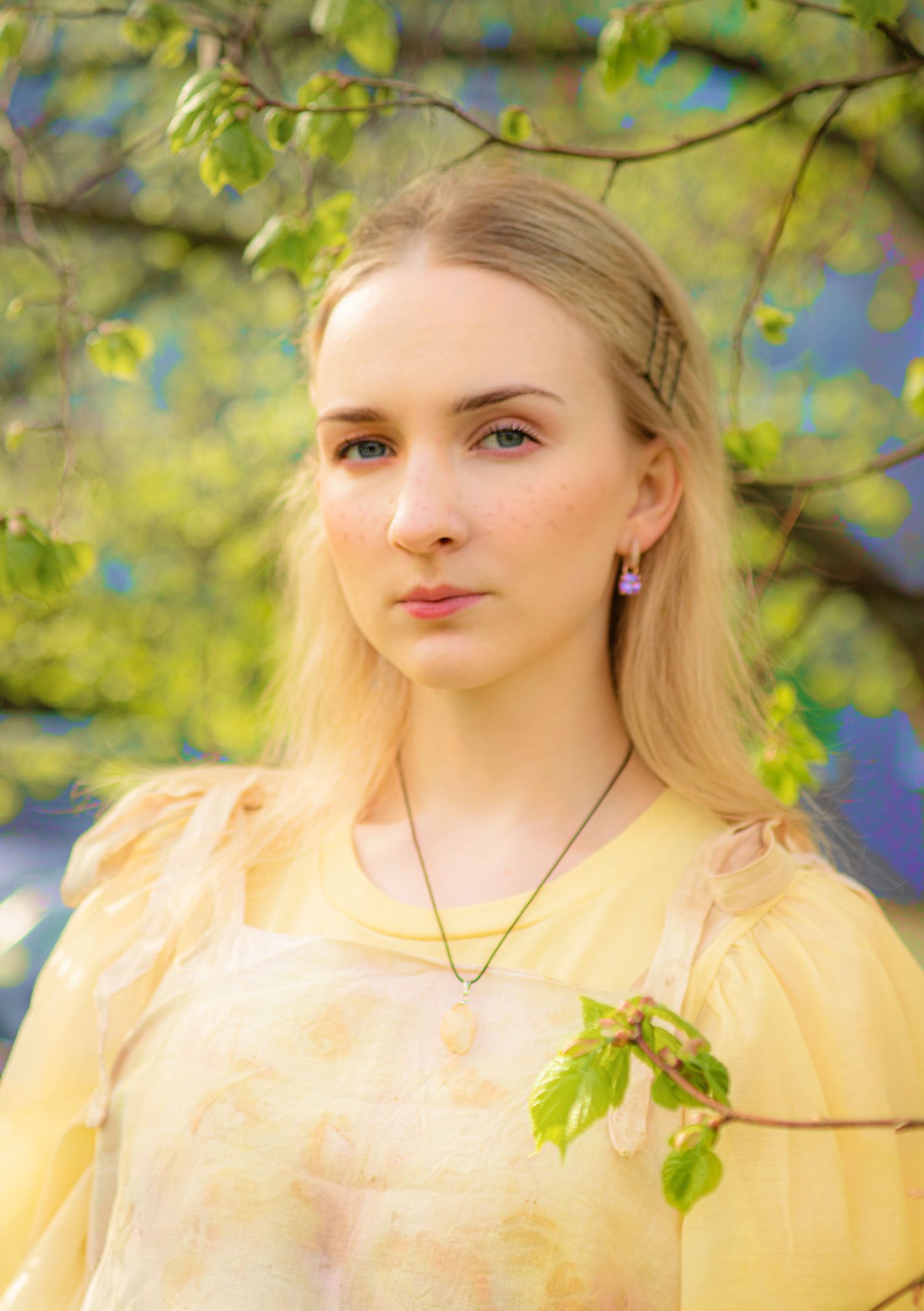 Young blonde woman in yellow dress with purple earrings and necklace standing near spring blossoms.