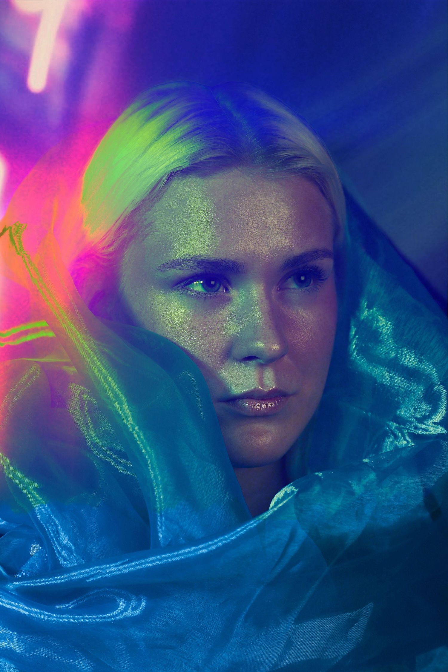 Blonde woman wrapped in teal fabric under vibrant neon purple and green lights in artistic portrait.