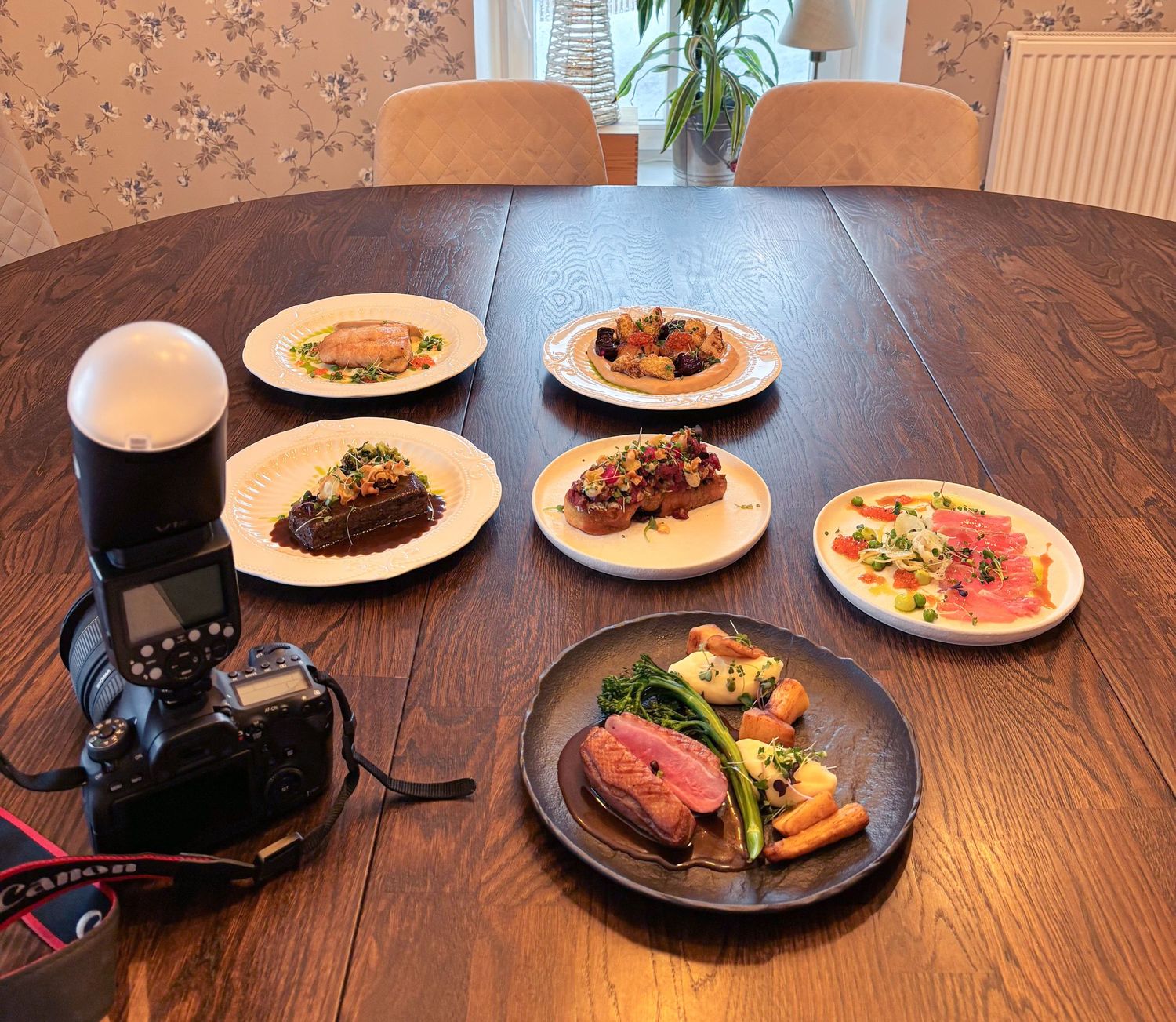 A DSLR camera with flash photographs five gourmet meat dishes on white plates arranged on a dark wooden dining table.