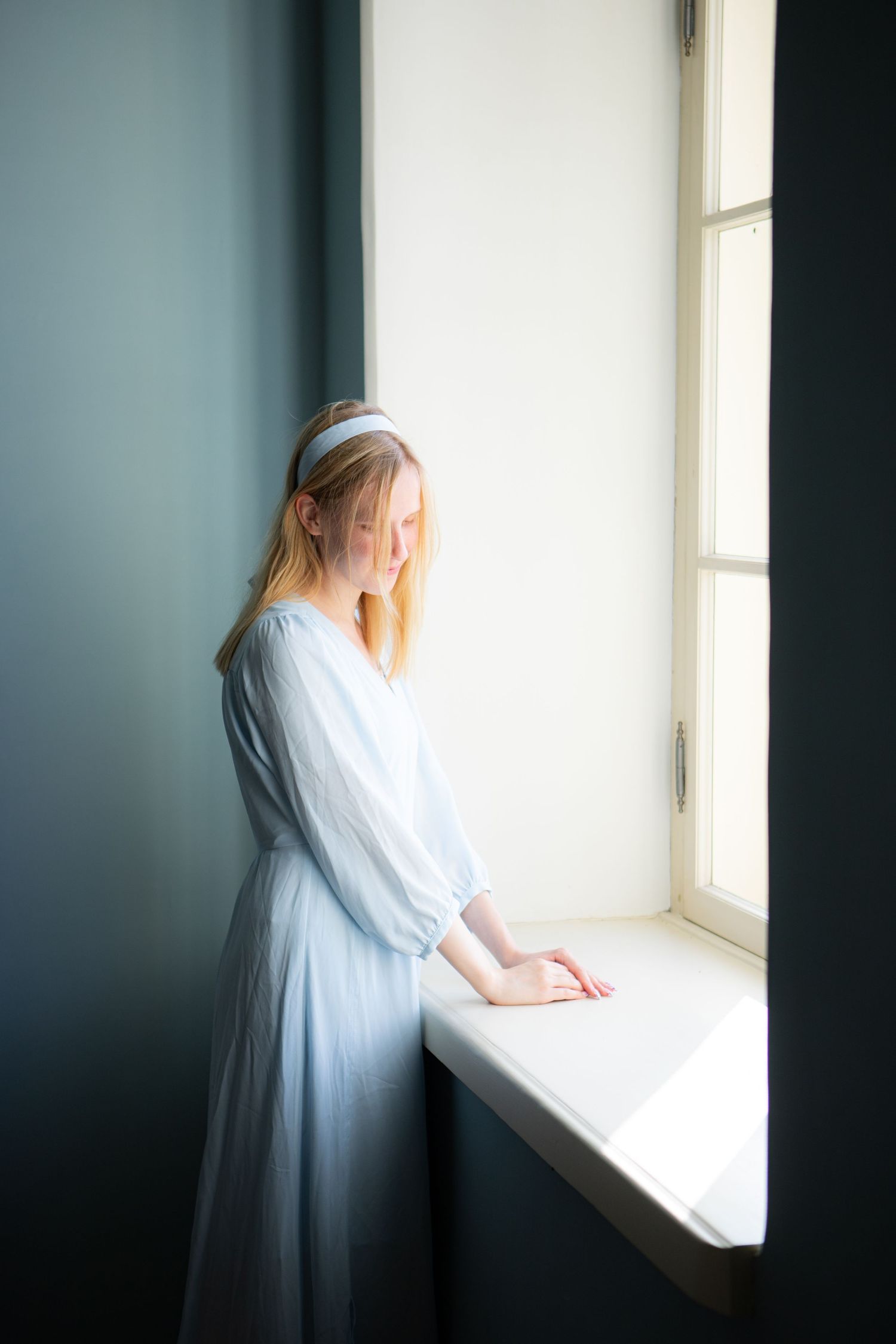 Young blonde woman in light blue dress standing by a bright window in a softly lit room.