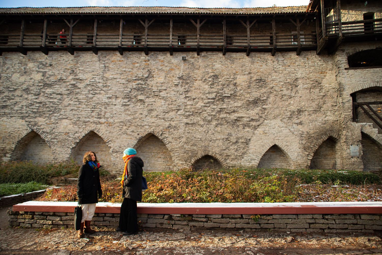 Two women walk along a medieval stone castle wall with arched niches in autumn.
