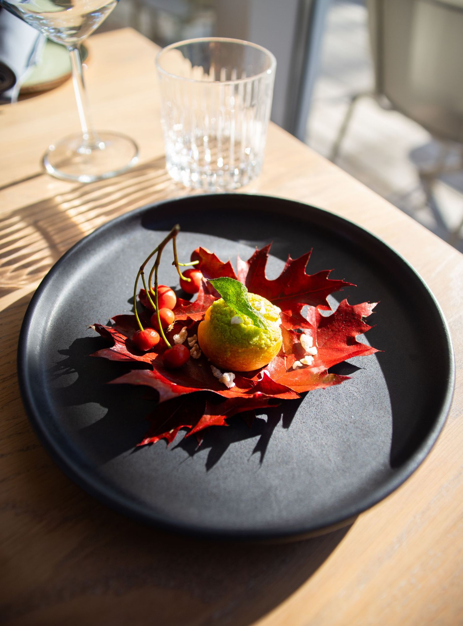 Artistic restaurant dish with green sorbet on red maple leaf with berries on black plate.