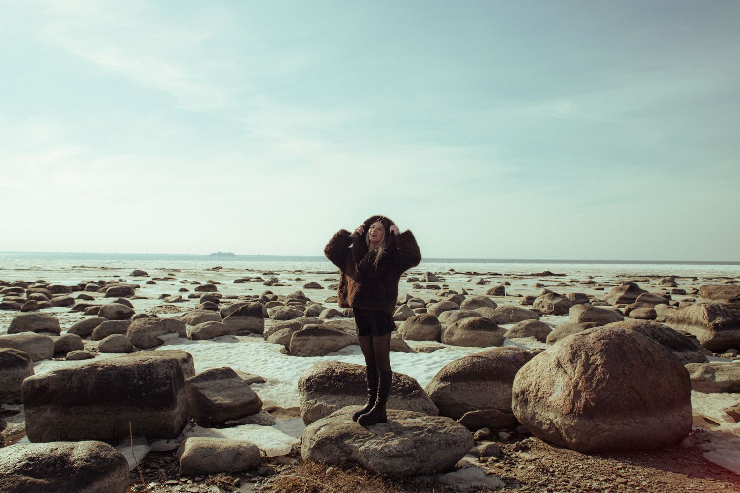 A woman in black stands on coastal rocks by the ocean under a blue sky.
