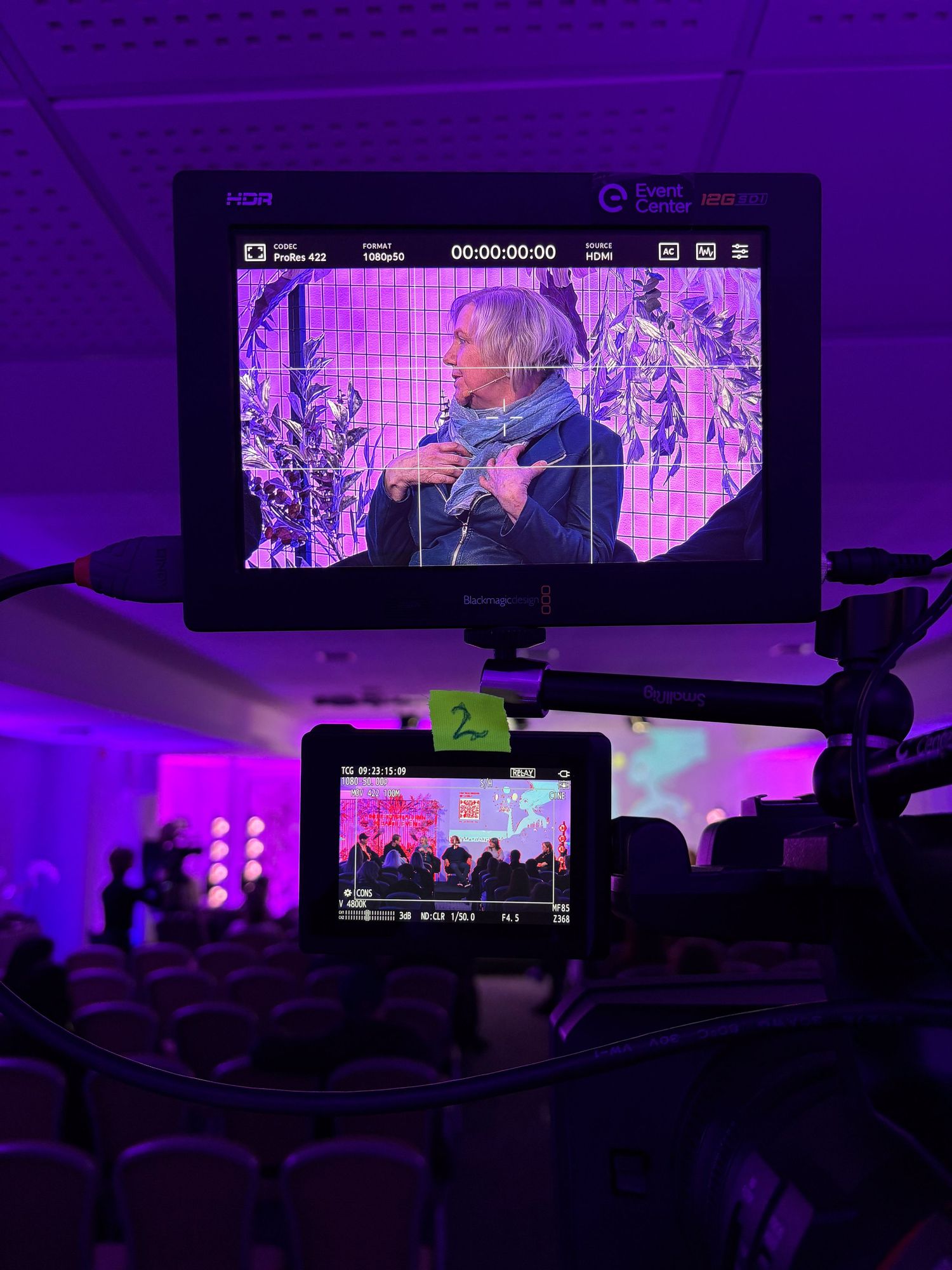 A camera operator films a woman speaking on stage at a purple-lit event venue through a monitor viewfinder.