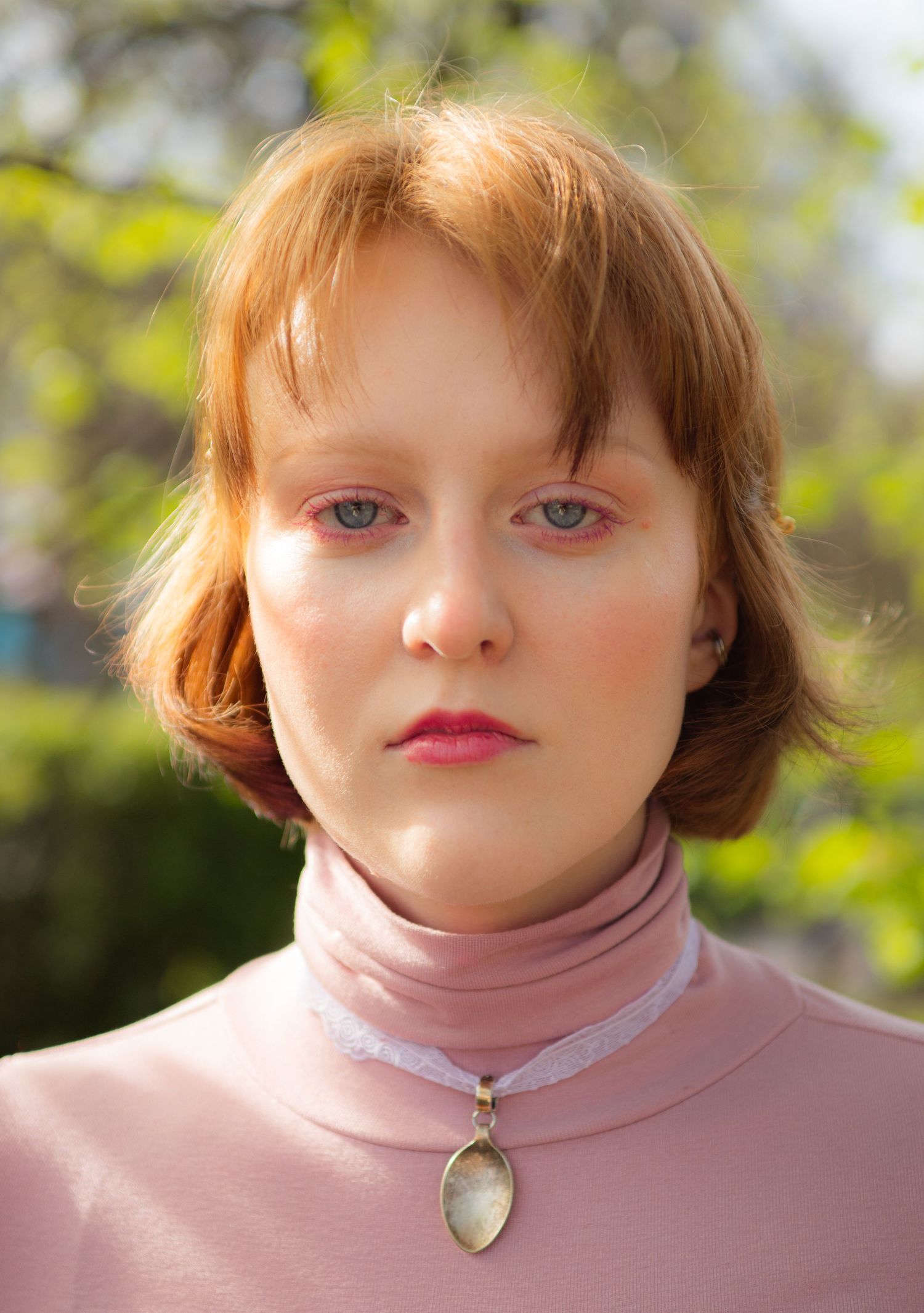 Close-up portrait of a young redhead woman in a pink turtleneck with a crystal pendant necklace.