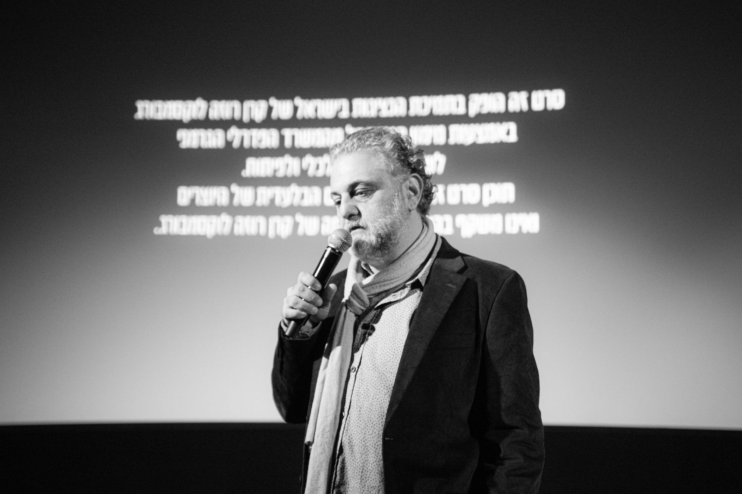 Black and white photo of a man speaking into microphone with Hebrew text projected behind.