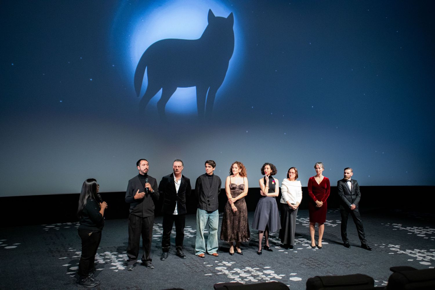 Film cast and crew lineup on stage at a movie premiere with a glowing wolf silhouette projected behind them.