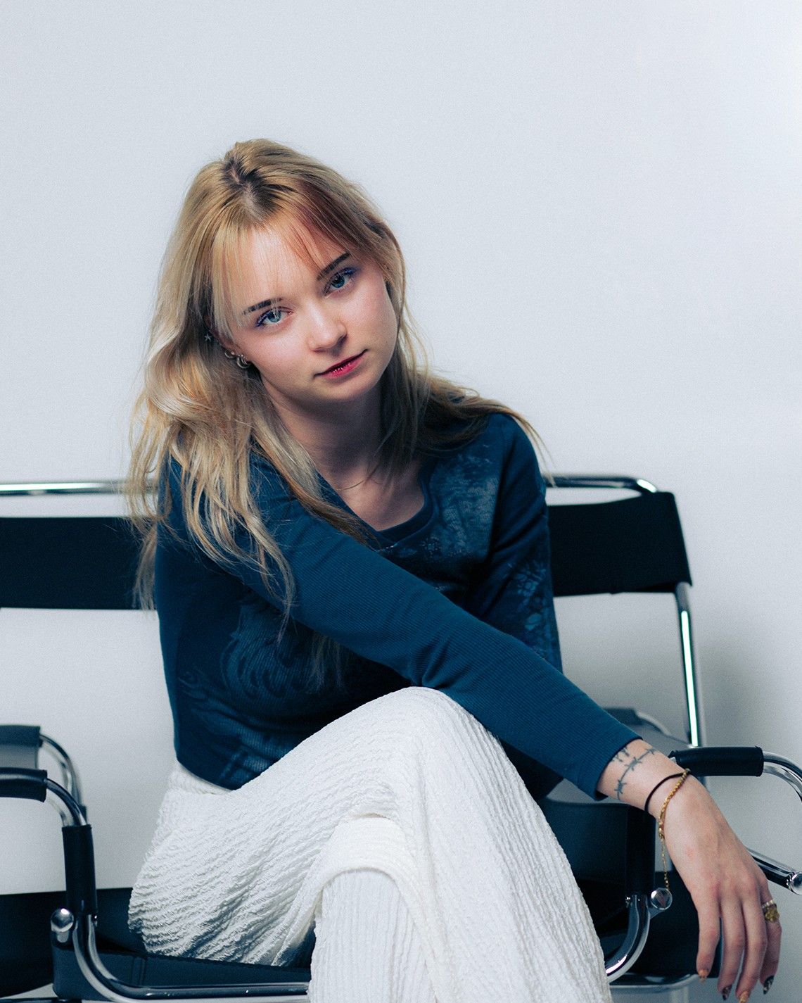 Young blonde woman in blue top and white pants sitting relaxed on a modern chrome chair against white background.