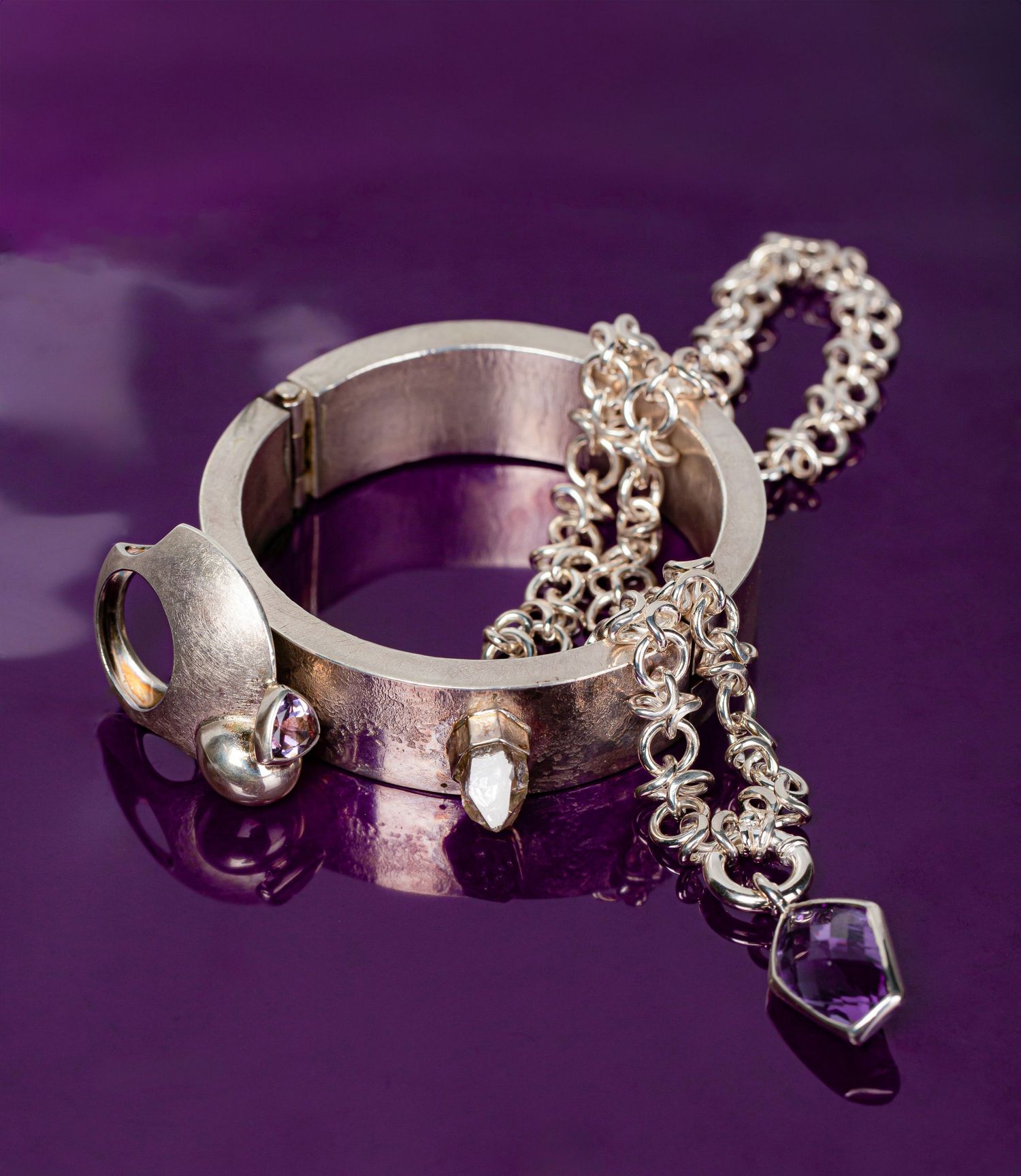 Silver bangle bracelet with chain and amethyst crystal pendant on purple reflective surface.