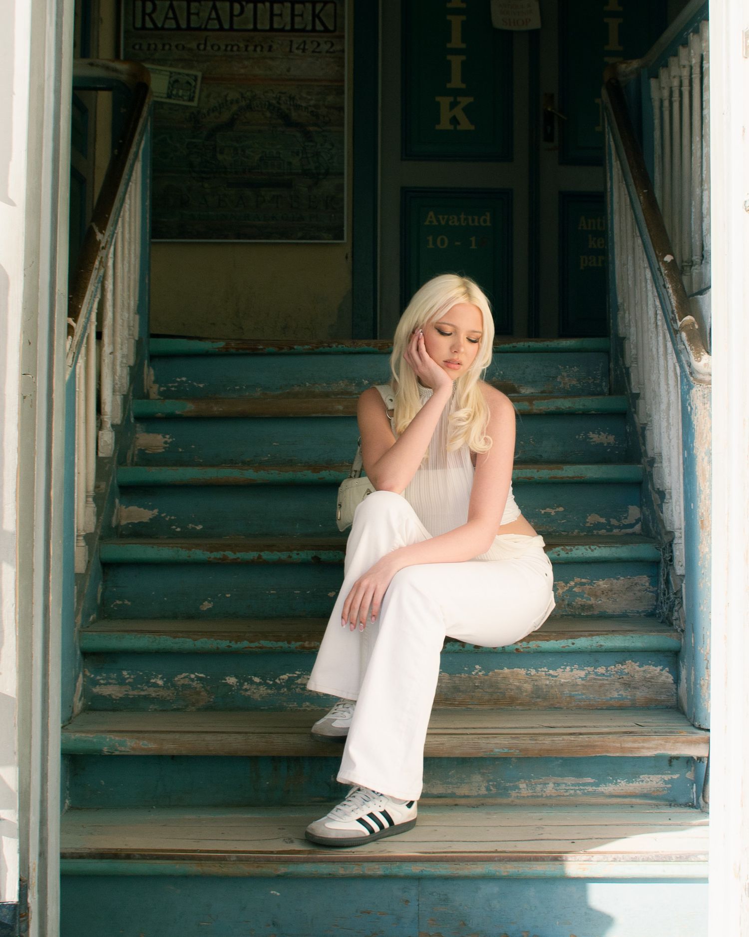 Blonde woman in white outfit and Adidas sneakers sitting pensively on teal painted wooden steps of vintage storefront.