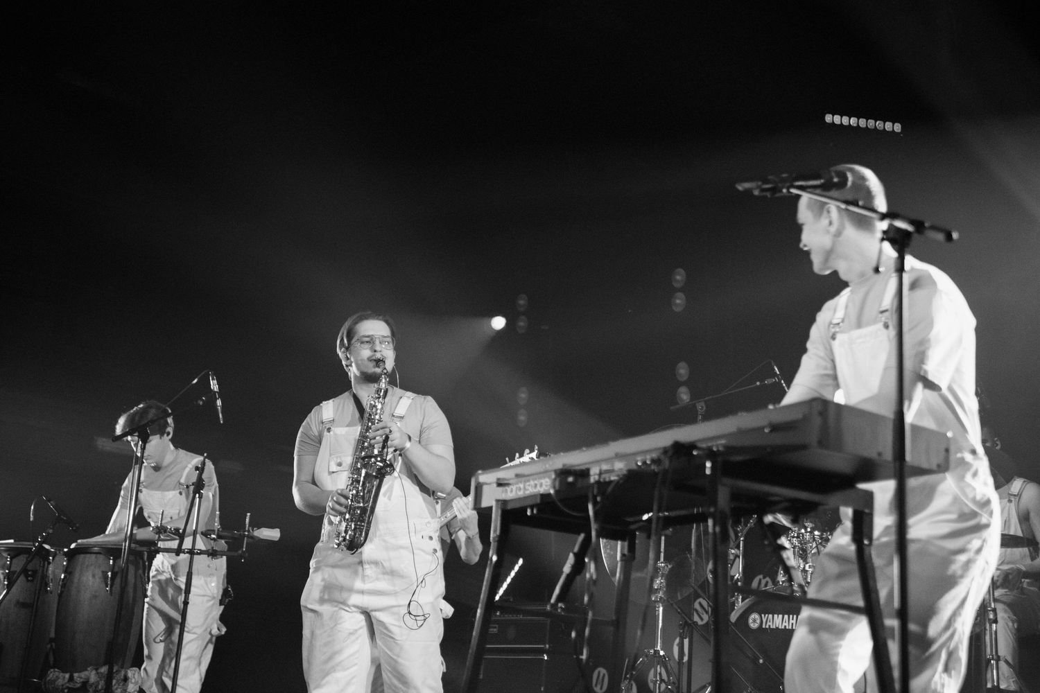 Black and white photo of a band performing on stage, featuring a saxophonist in white overalls.