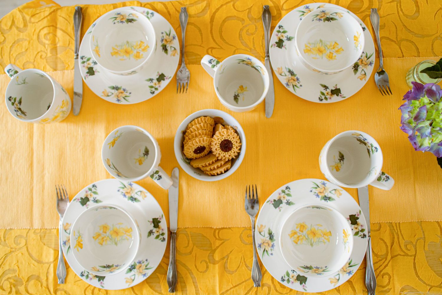 Overhead view of a yellow table set with floral white china dishes, cutlery, and a bowl of crackers.