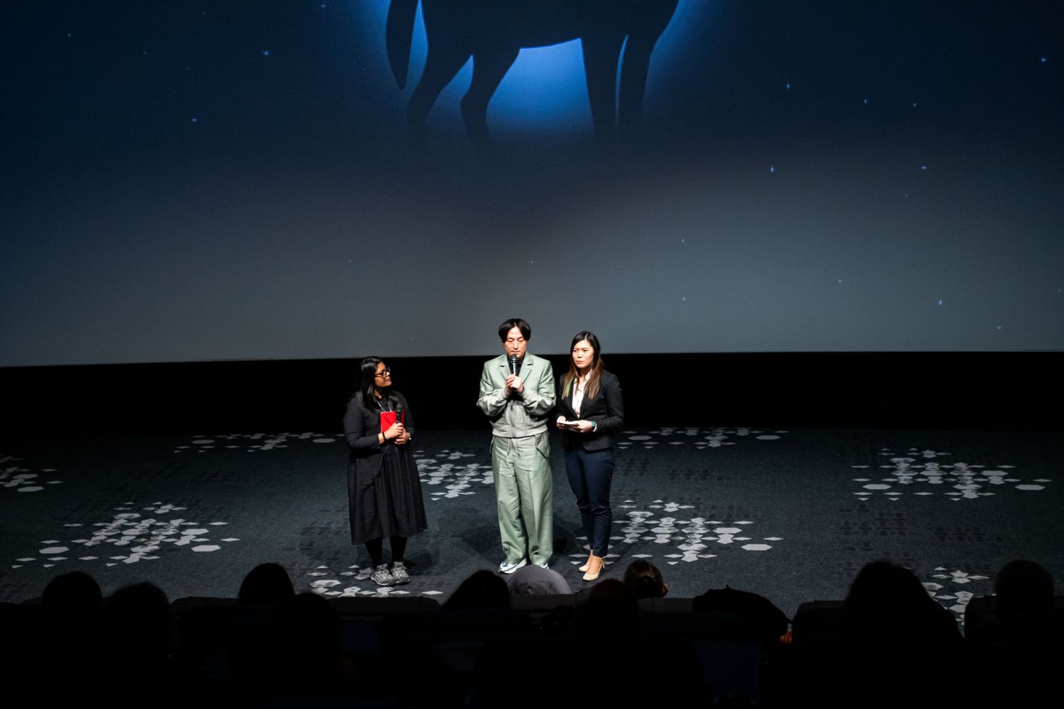 Three presenters standing on a decorated stage floor at an indoor film festival screening.